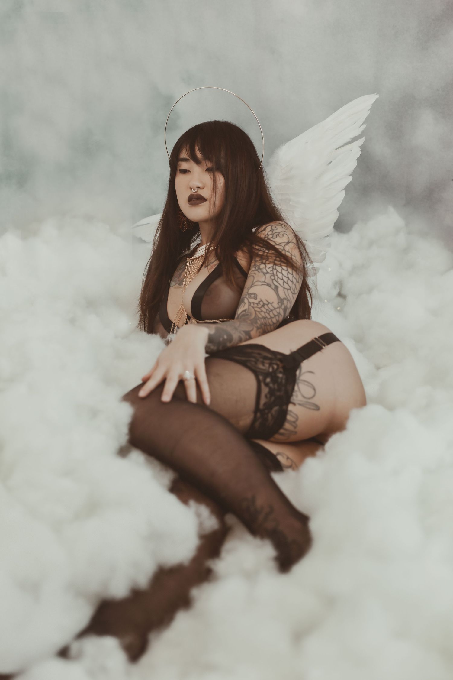 Dark artistic boudoir photo with angelic wings in misty clouds with gothic lingerie and tattoo aesthetic.