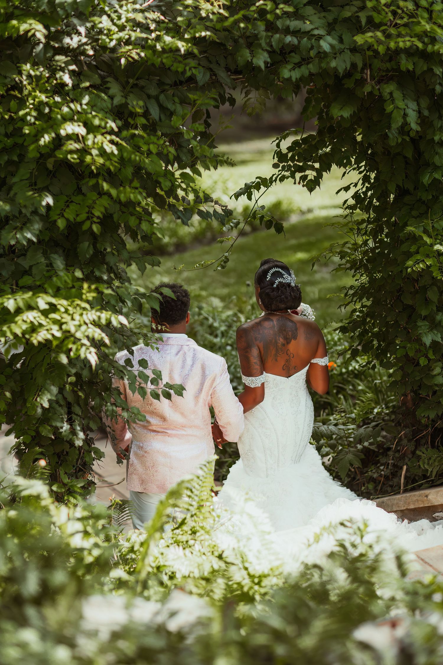 Couple in wedding attire walks through garden path framed by green leaves.