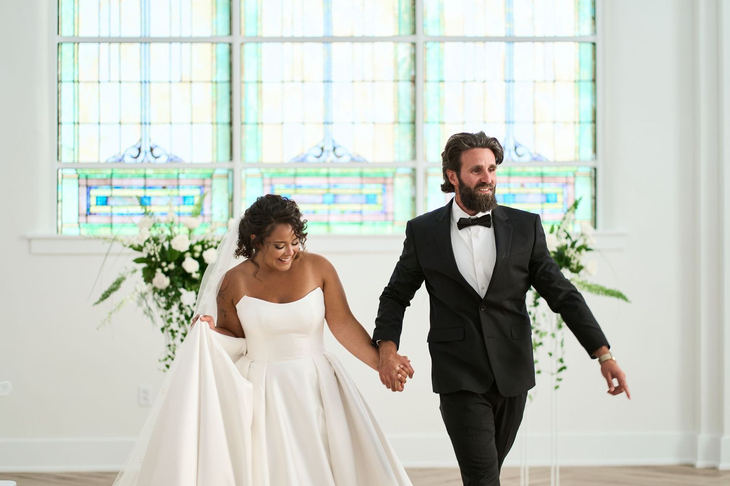 A joyful newlywed couple walking hand in hand in a bright, airy chapel with stained glass windows.