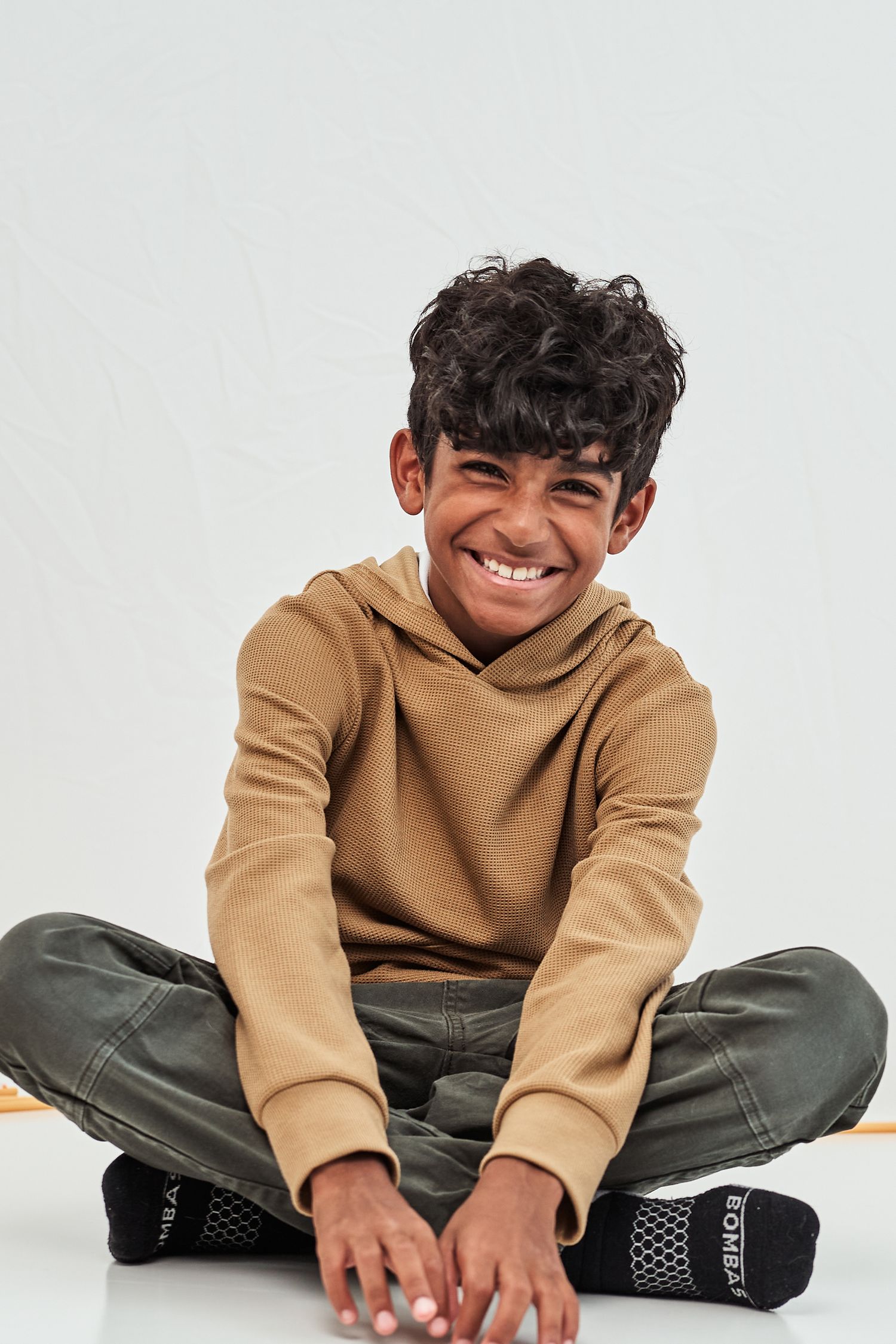 A figure in a tan hoodie and olive pants sits cross-legged on a white background with a playful expression.