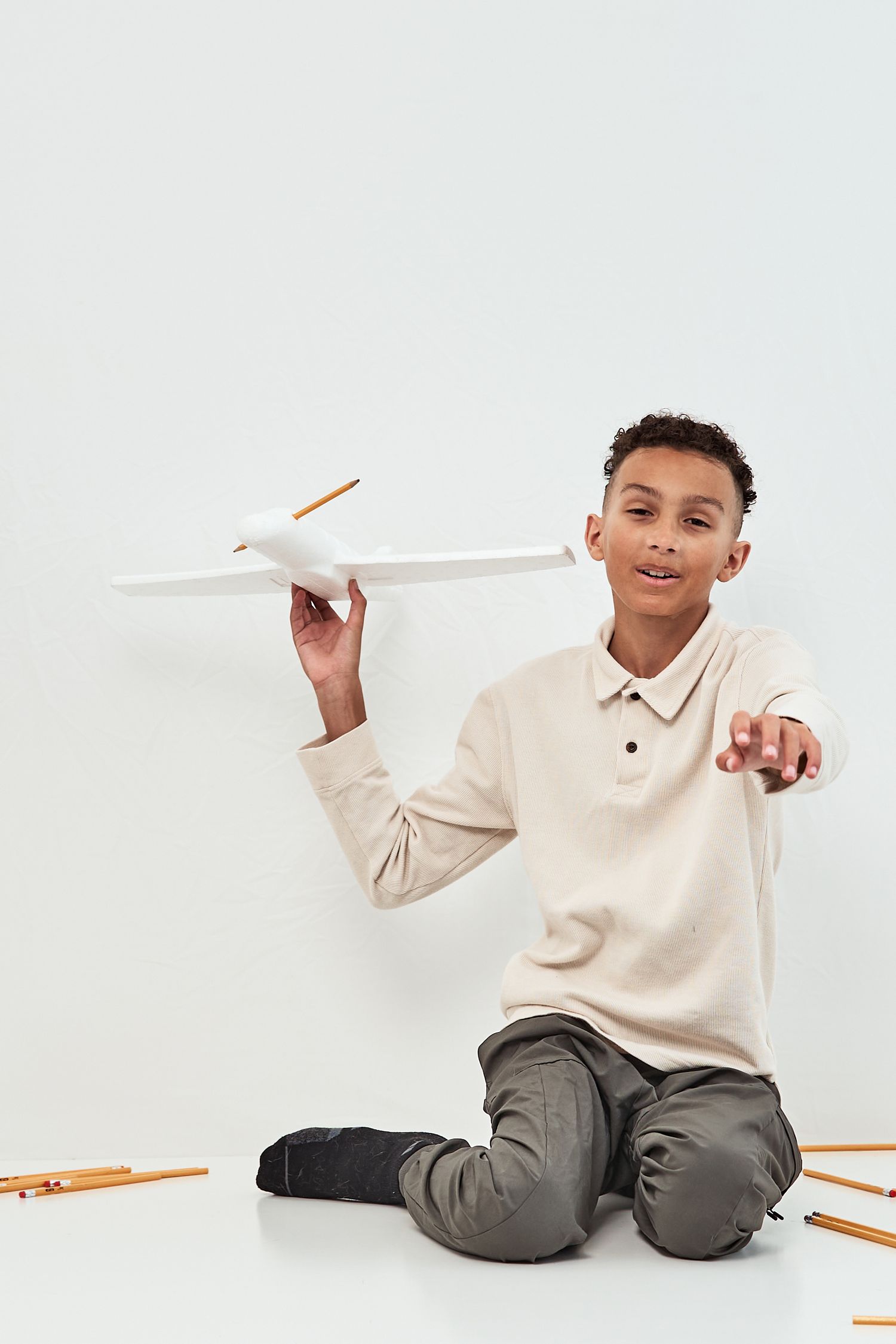 Young person in beige sweater sits on floor playing with paper airplane against white background.