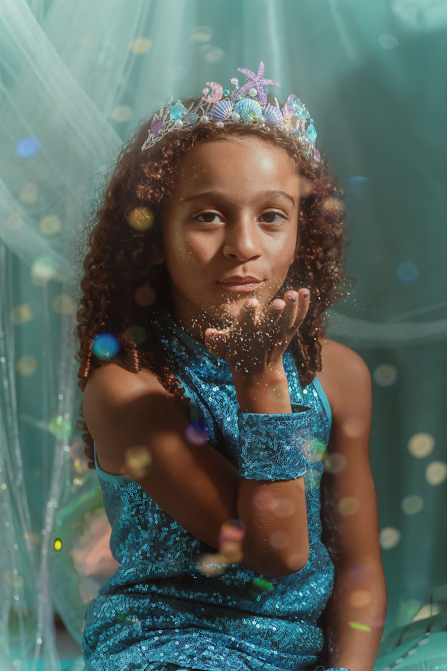 A young girl in a blue sequin dress and mermaid crown blows glitter from her hand against a teal backdrop.