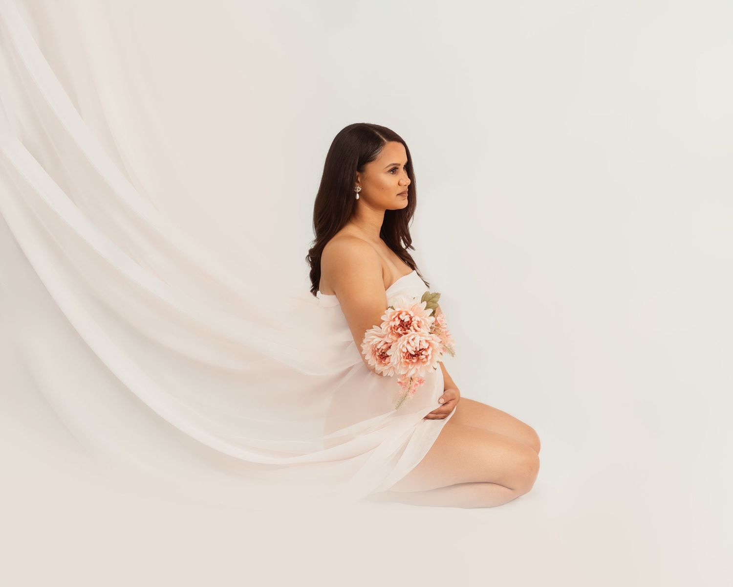 Maternity portrait with flowing white fabric and pink floral bouquet against light backdrop.