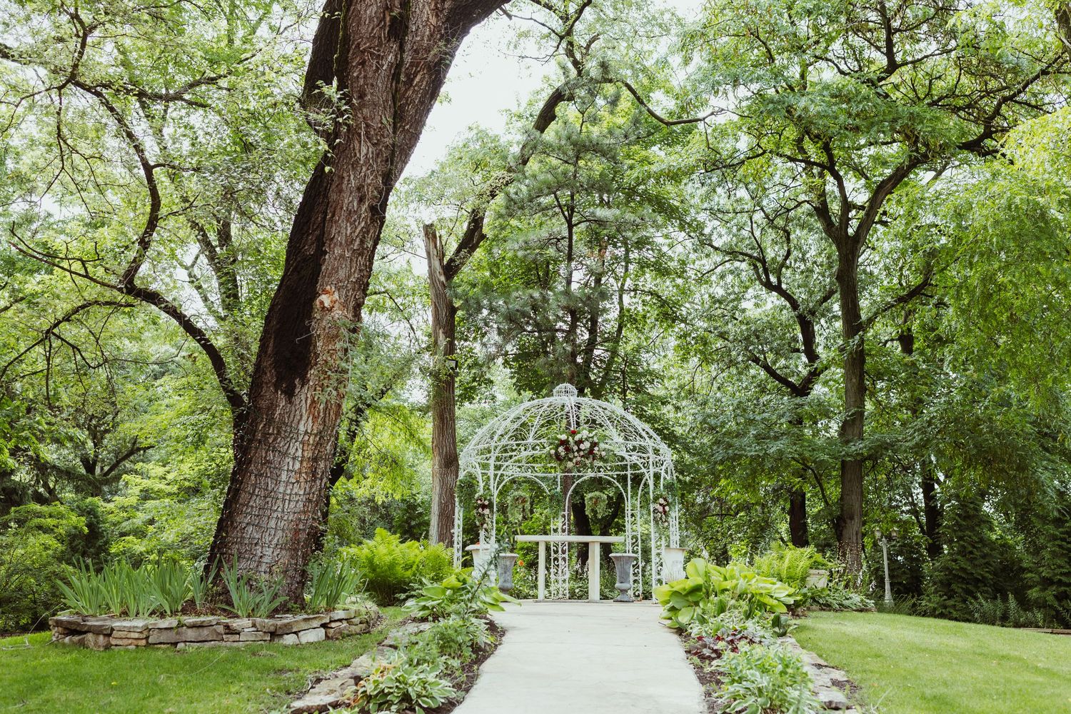 A white garden gazebo with stone pathway surrounded by trees and landscaping.