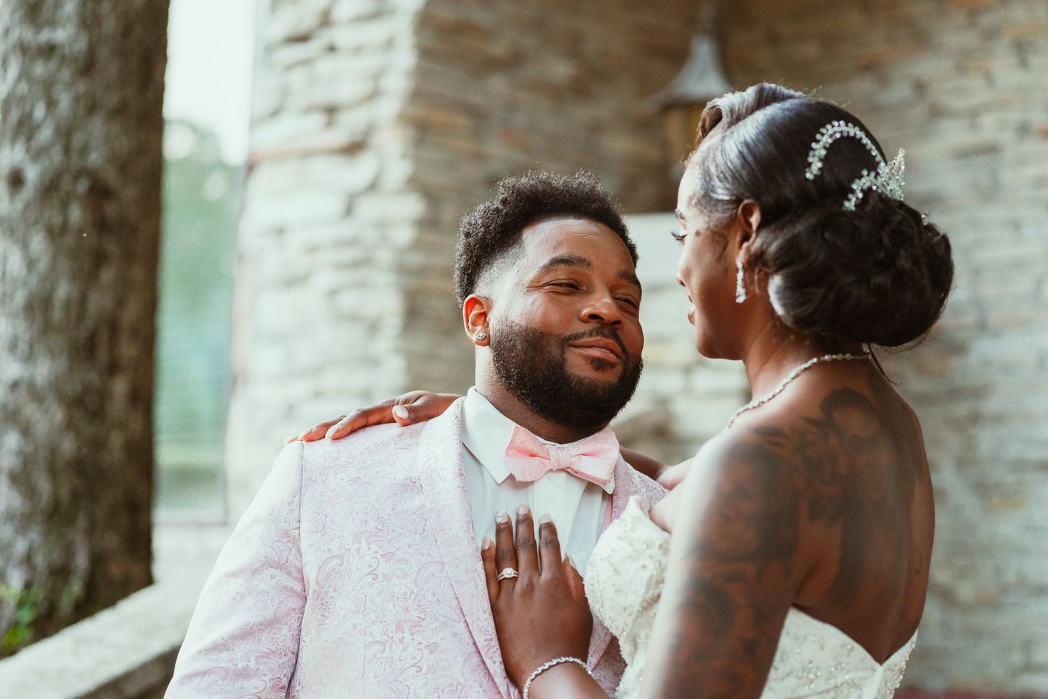Wedding couple in white and pink attire share a tender moment against a stone wall background.