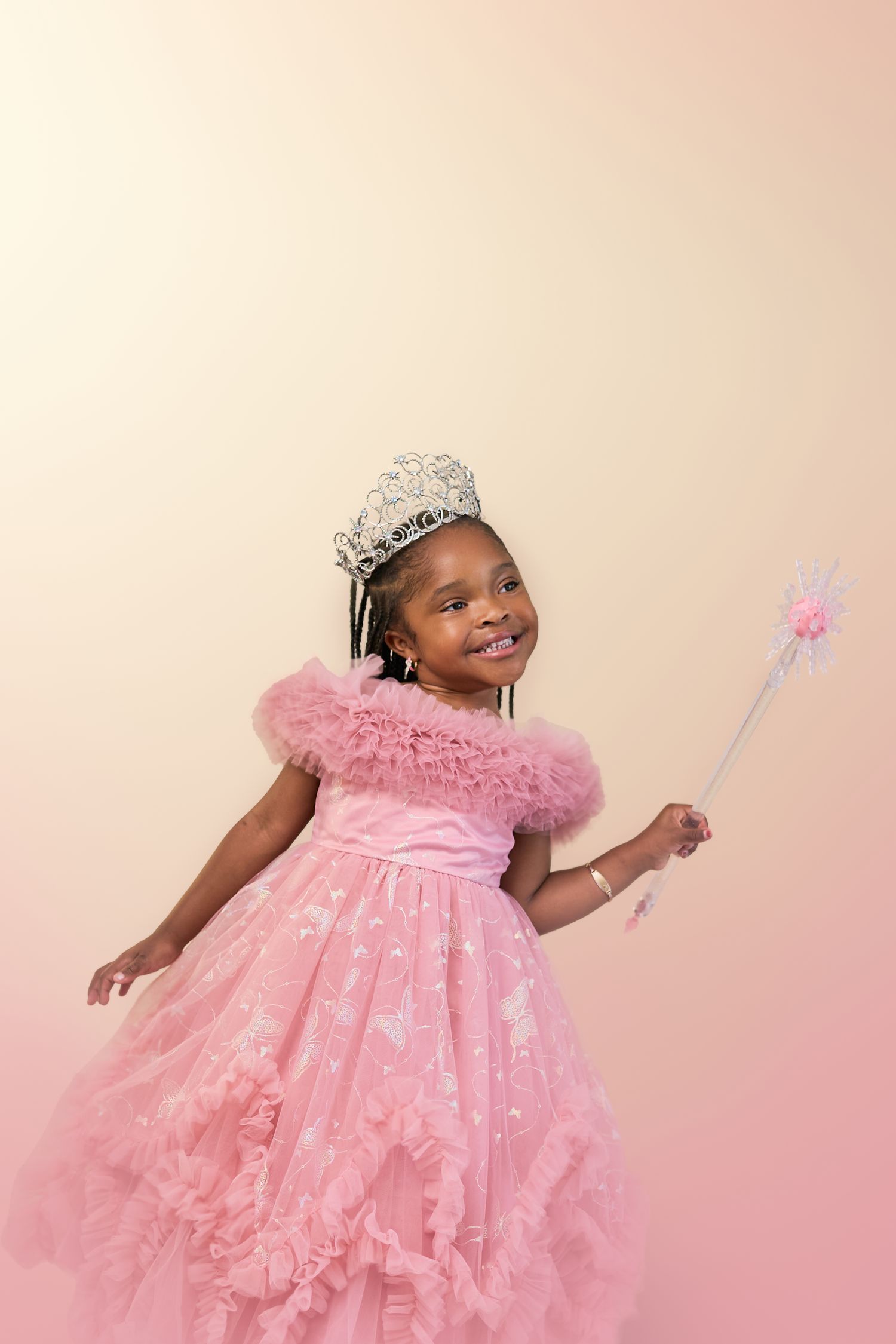 A young child wearing a pink princess dress and tiara dances joyfully against a soft pink studio backdrop.