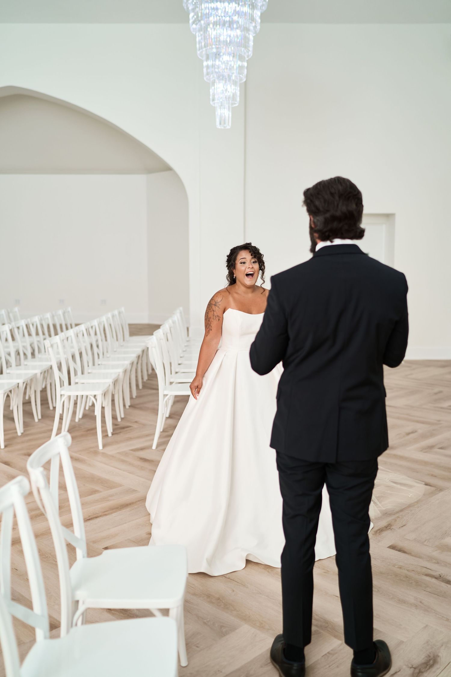 Wedding couple stands in elegant white chapel with rows of chairs during their first look.
