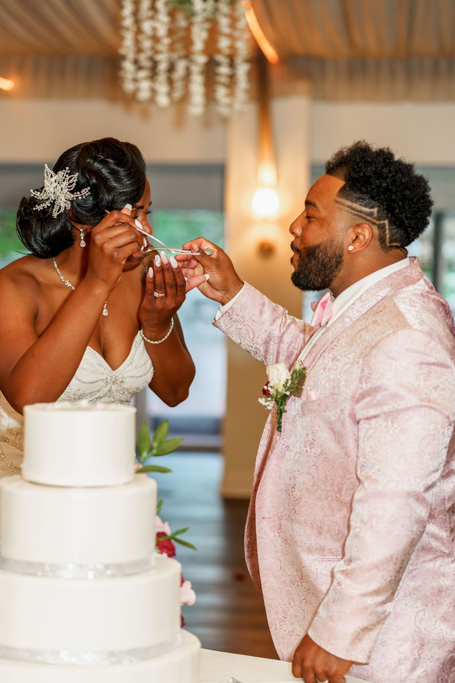 Playful cake-feeding moment between newlyweds beside their white tiered wedding cake.