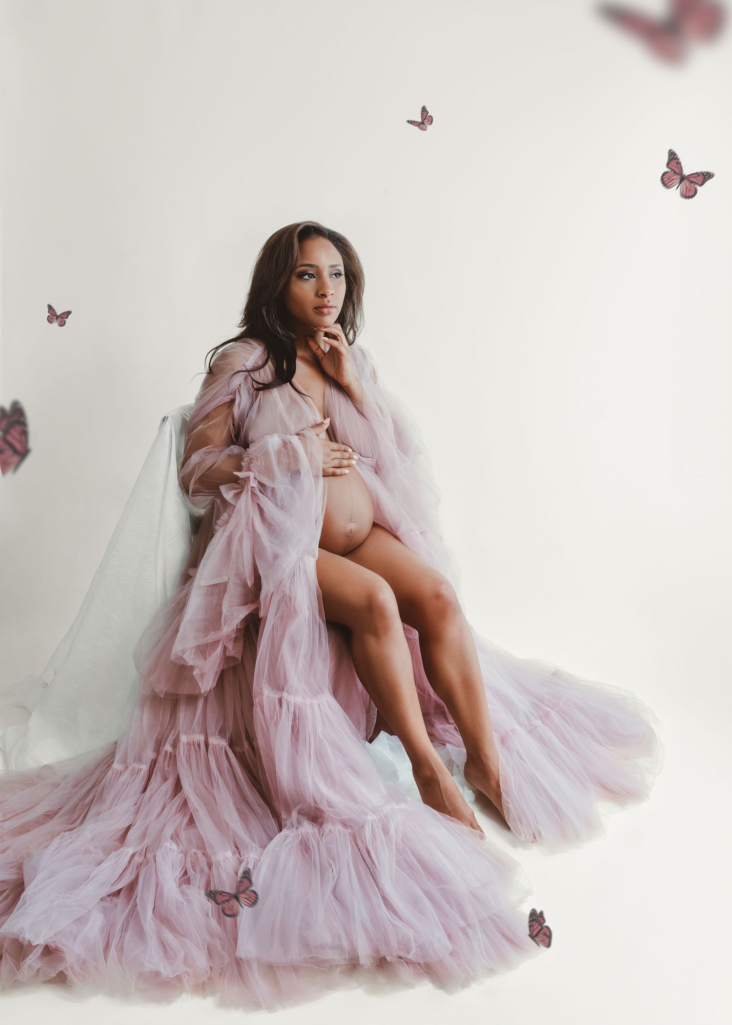 Maternity photo with flowing pink dress and floating butterflies against white backdrop.