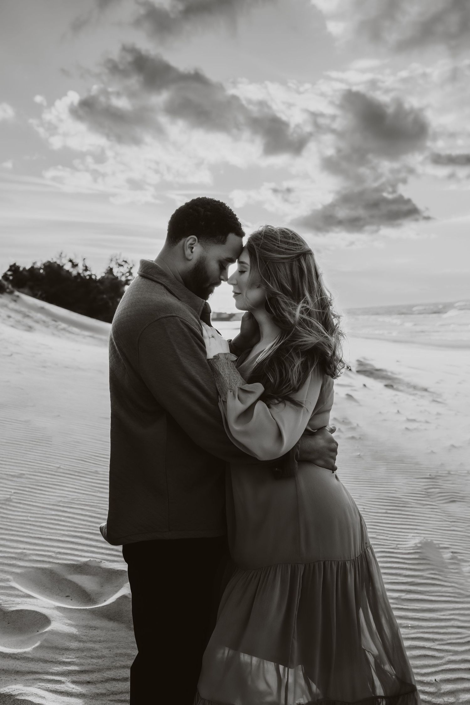 A romantic black and white portrait of a couple embracing on sandy dunes at sunset.