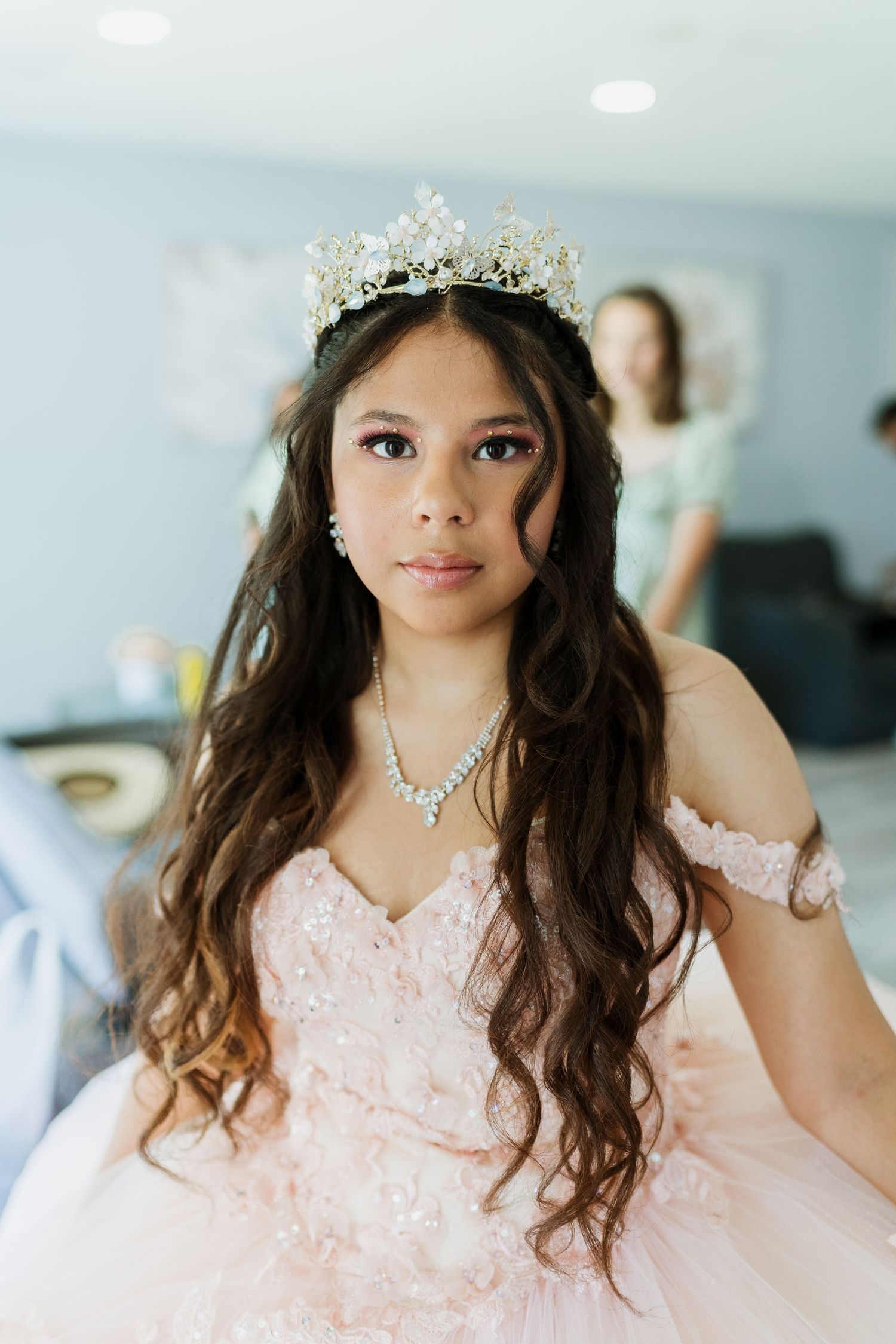 A sparkling tiara crowns flowing dark hair over a pink off-shoulder gown with delicate jewelry.