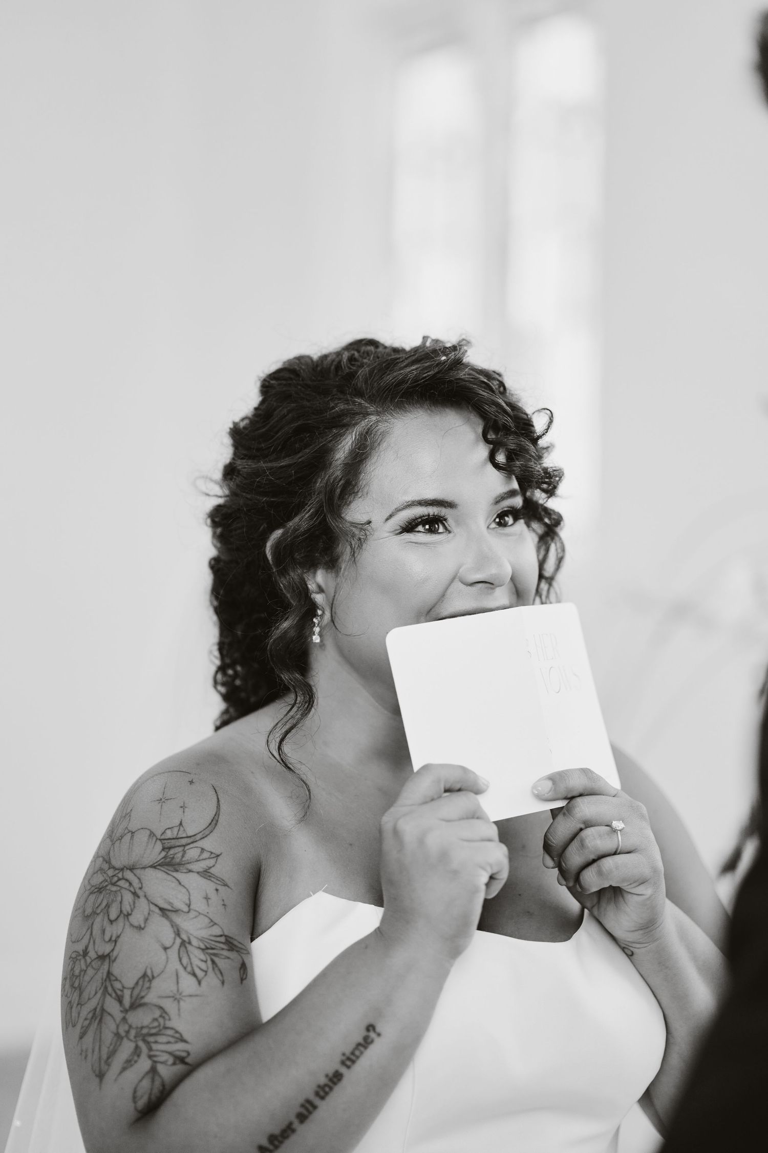 A bride in a white dress holds up a note while smiling, showing off her floral arm tattoo.