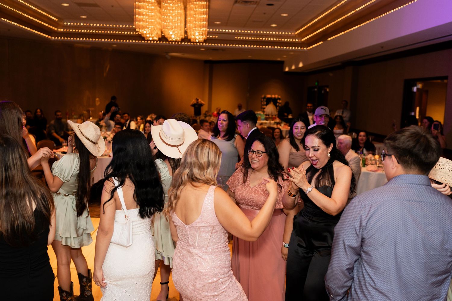 Wedding guests dance together on a dimly lit ballroom floor under elegant crystal chandeliers at an evening reception.