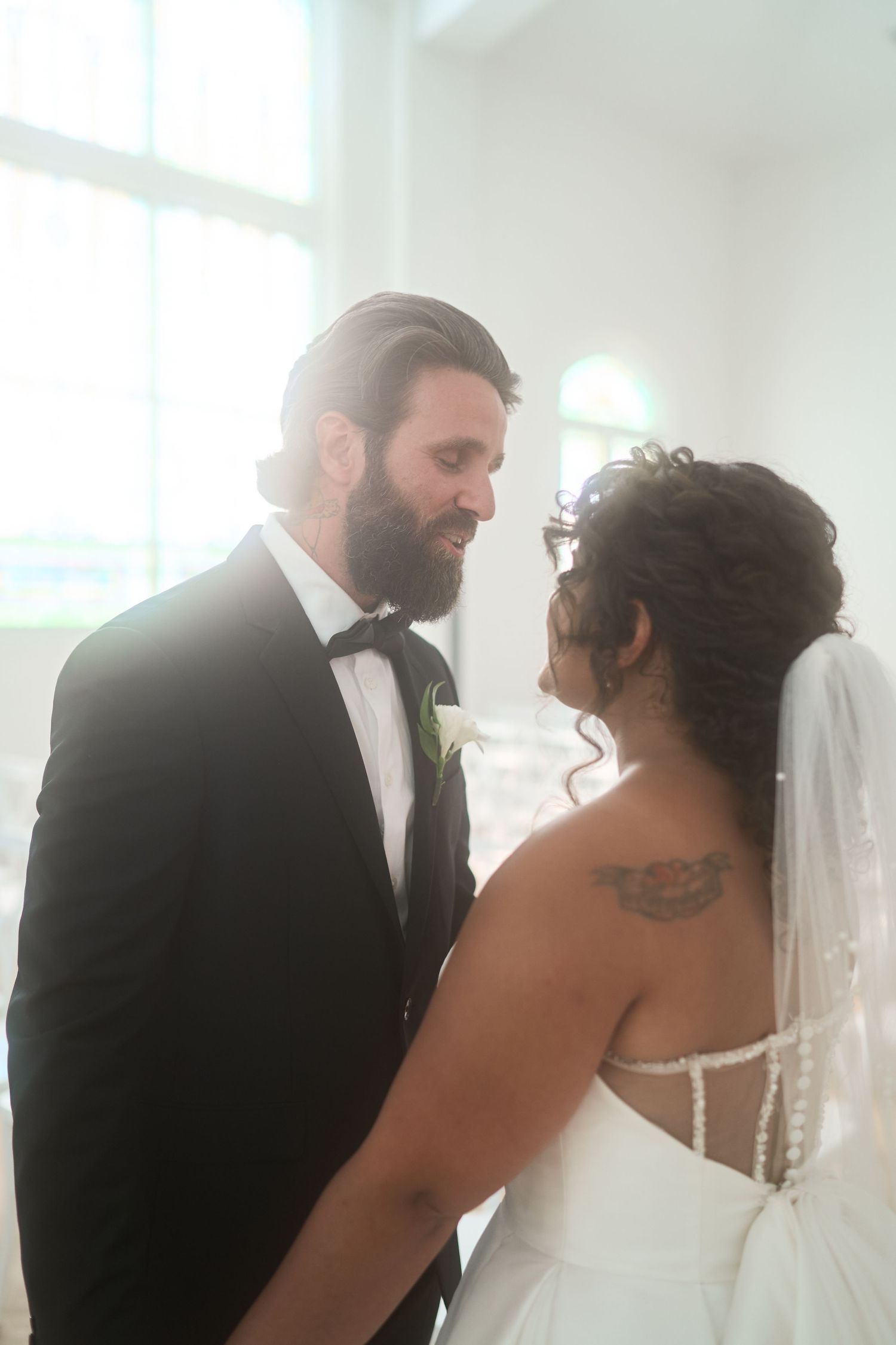 An intimate wedding moment in a sun-lit white chapel between bride and groom.