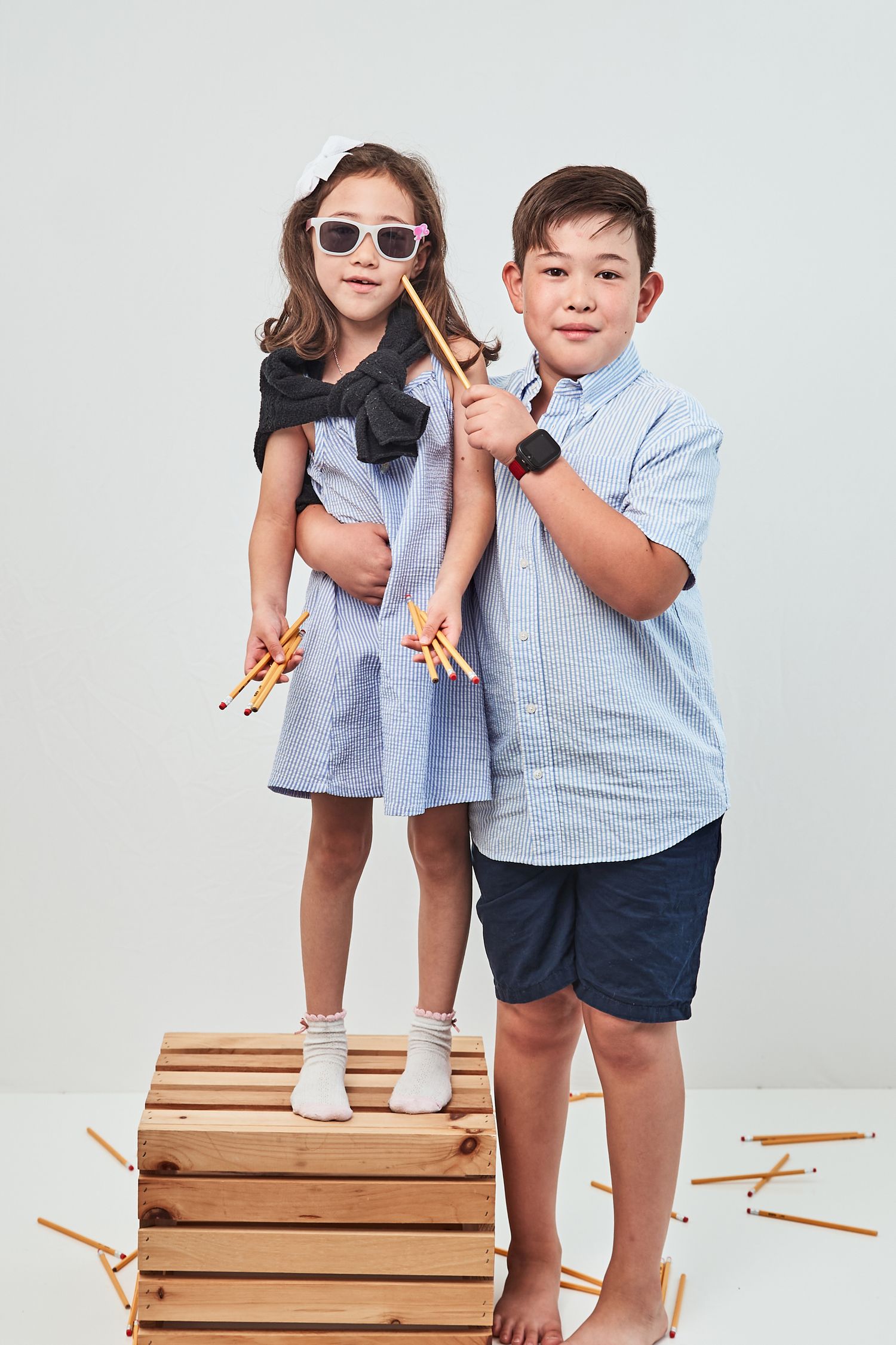 Two children standing by wooden crates holding slingshots, wearing summer casual clothing against white background.