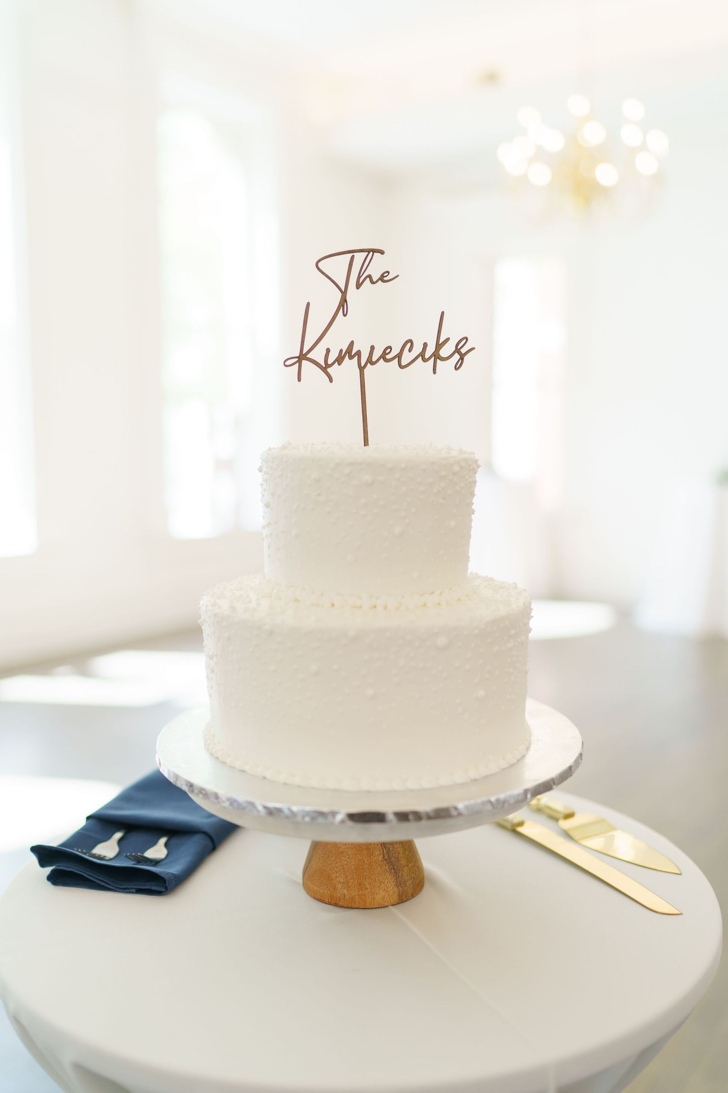 Simple two-tier white wedding cake with 'The Kupecks' topper on wooden stand with navy and gold accents.