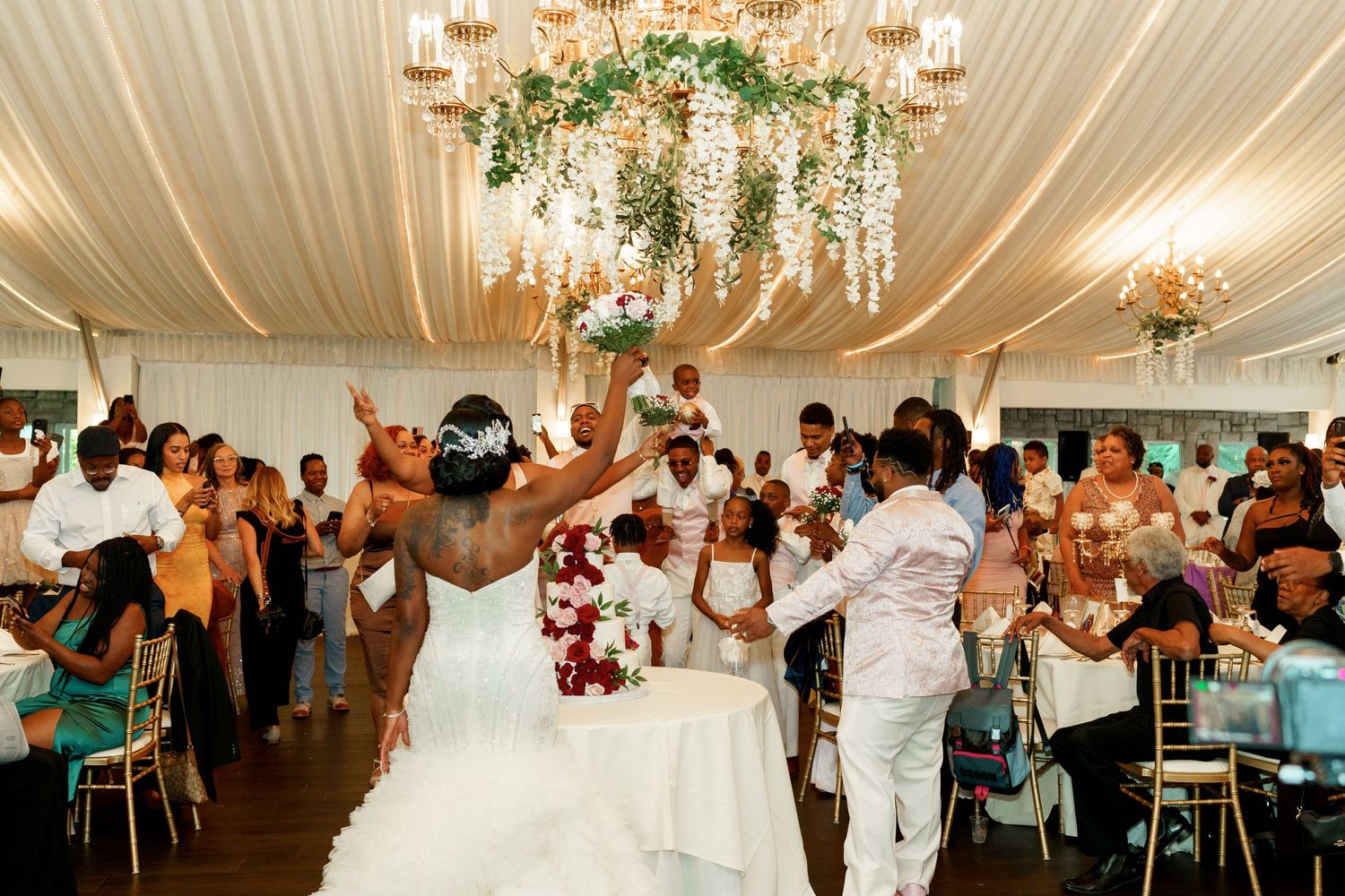 Jubilant wedding celebration with guests dancing beneath a draped ceiling and crystal chandelier.