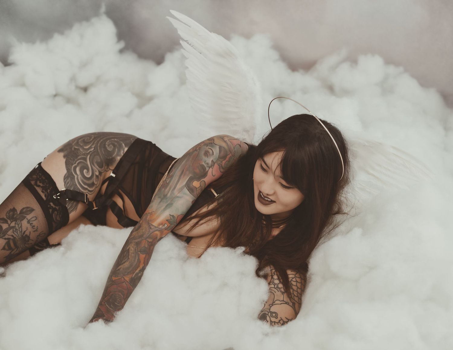 An artistic boudoir photograph featuring tattooed model posed among white cloud-like fabric.