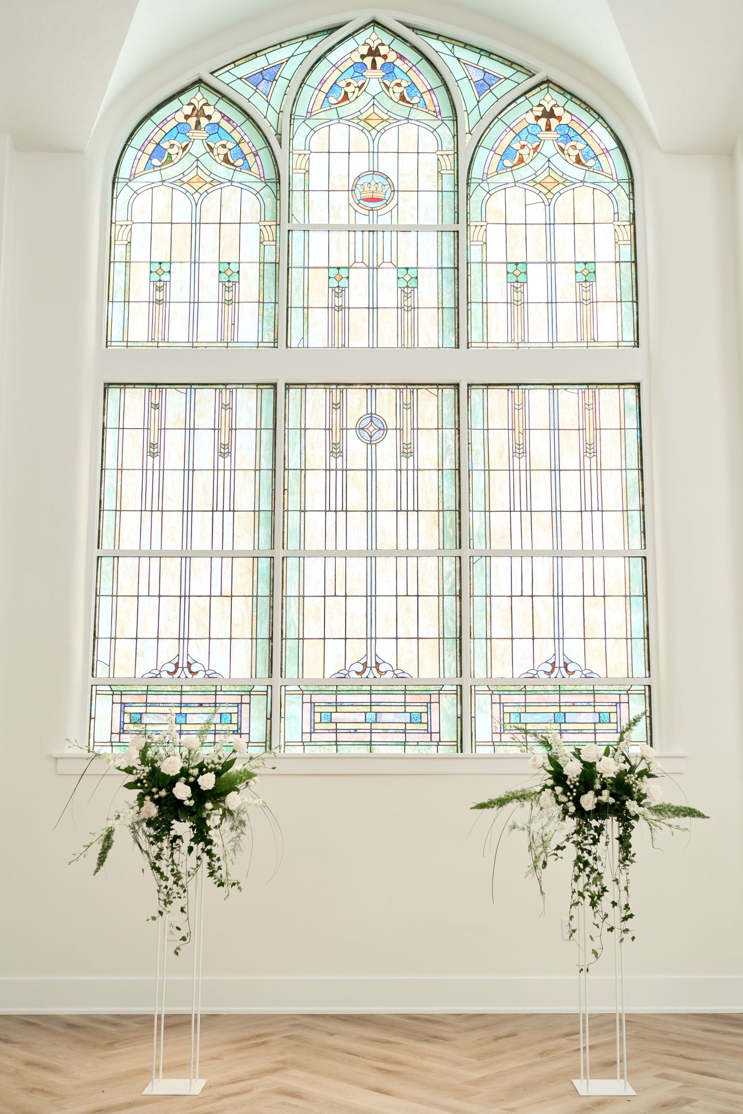 Elegant chapel venue with stained glass arched windows and white floral arrangements on columns.