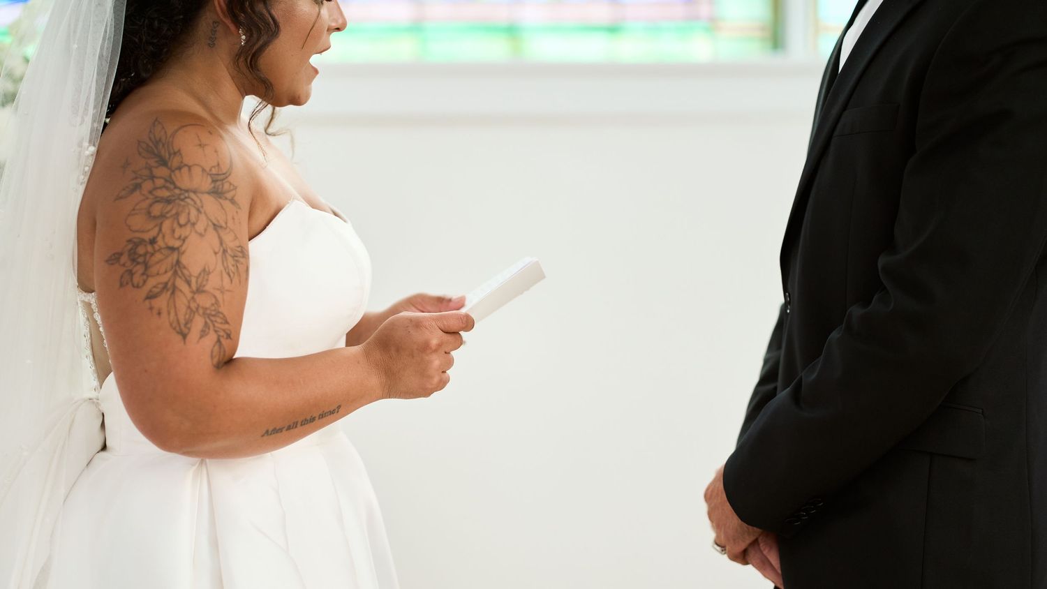 Two people in wedding attire face each other in a bright white room during a ceremony.