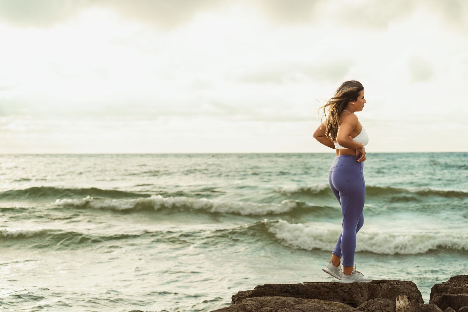 Woman in athletic wear stands on rocky outcrop overlooking ocean waves.