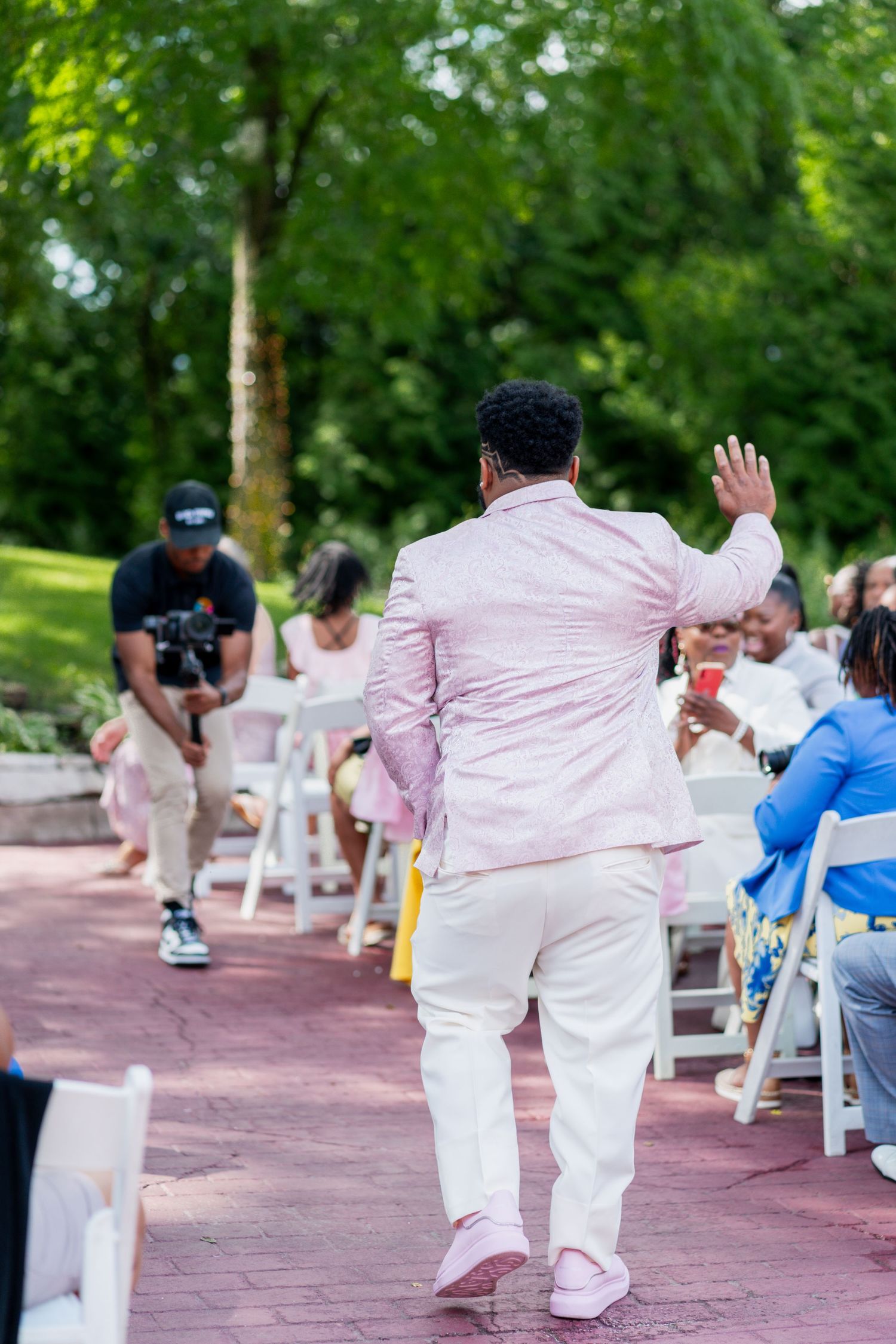 Groom in pink suit walks confidently down aisle at outdoor wedding ceremony.