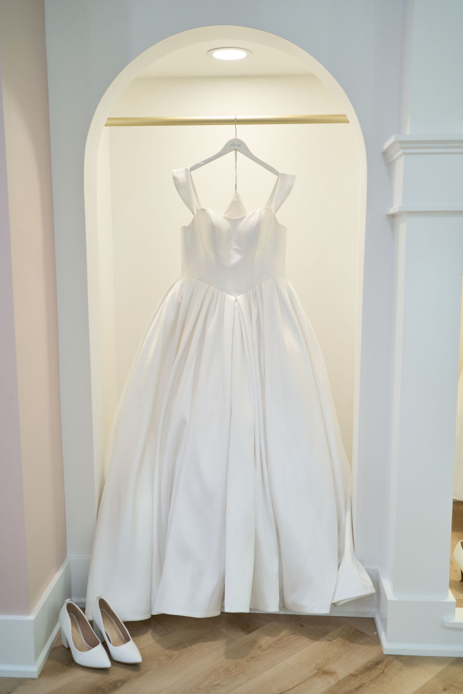 An elegant white wedding dress hangs in an arched alcove with matching shoes below.