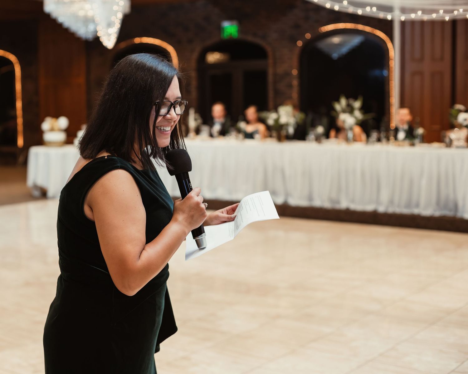 A person in black reads from notes while standing in an elegant reception venue.