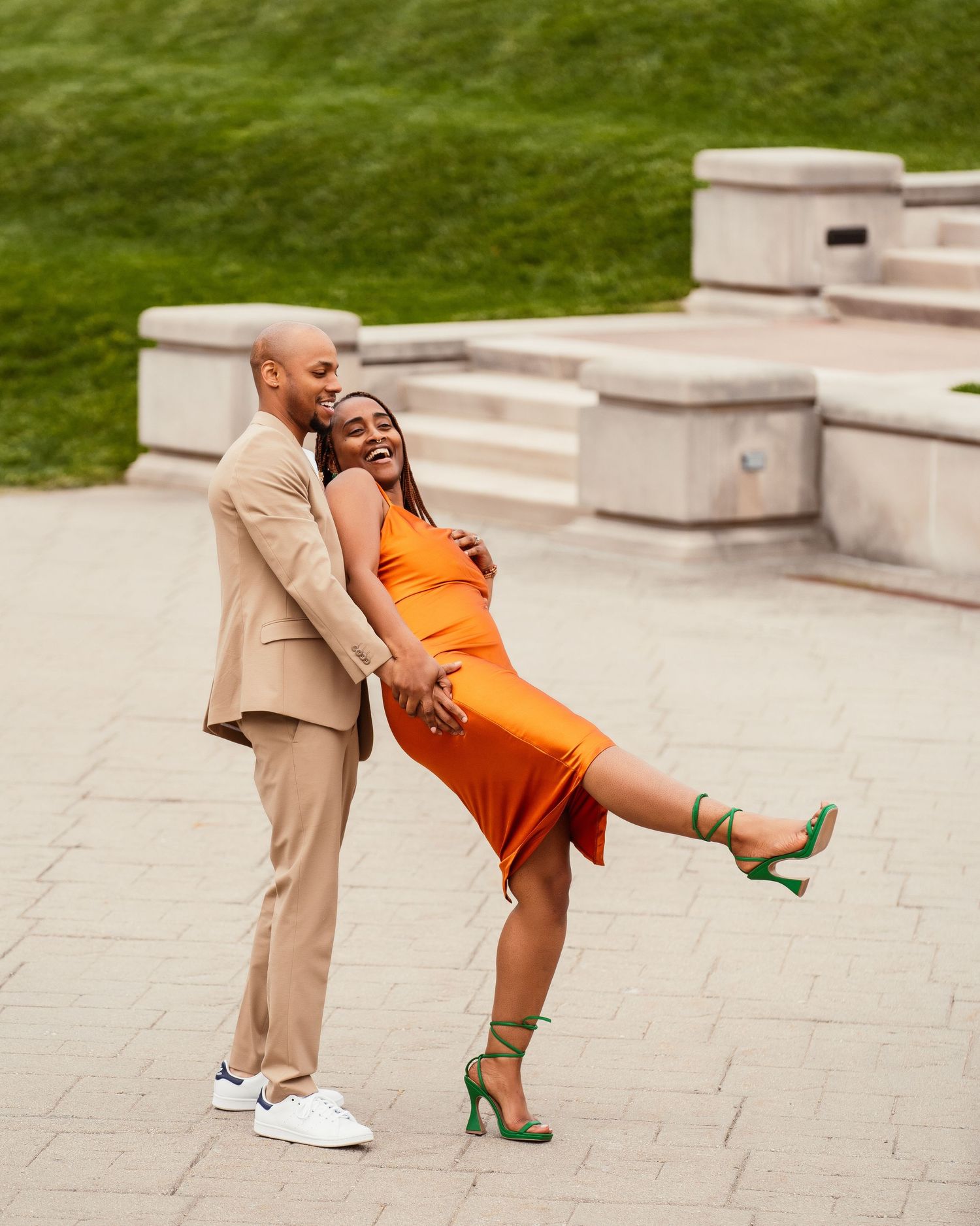 Playful couple in orange dress and beige suit dancing and laughing on concrete steps.