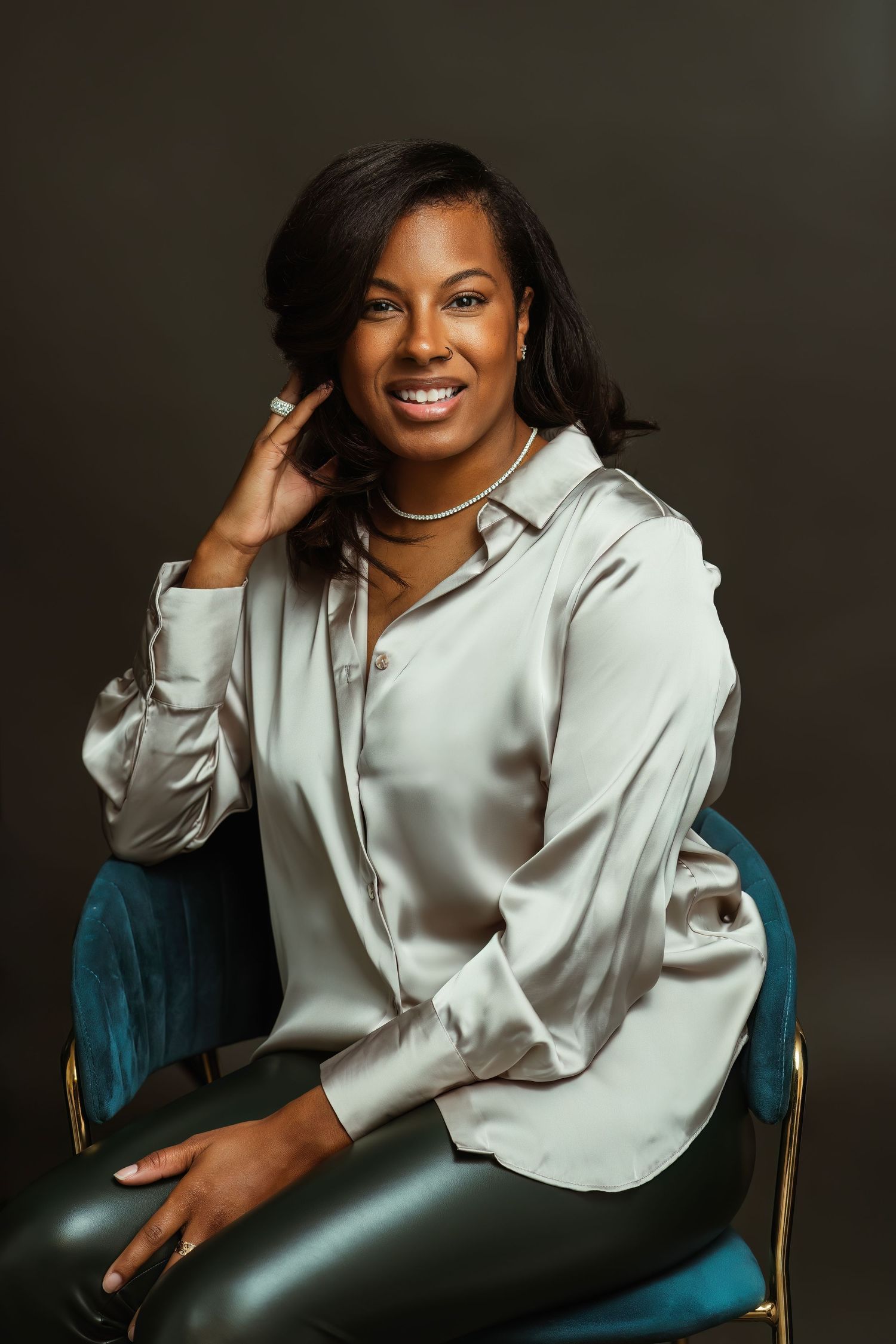 Professional studio portrait of someone in a white blouse and leather pants posing elegantly on a teal velvet chair.