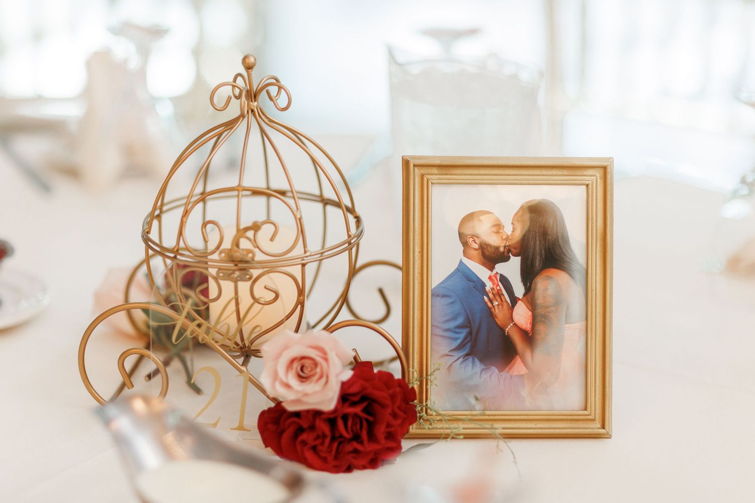 Decorative gold birdcage with pink and red roses next to framed photograph on table setting.