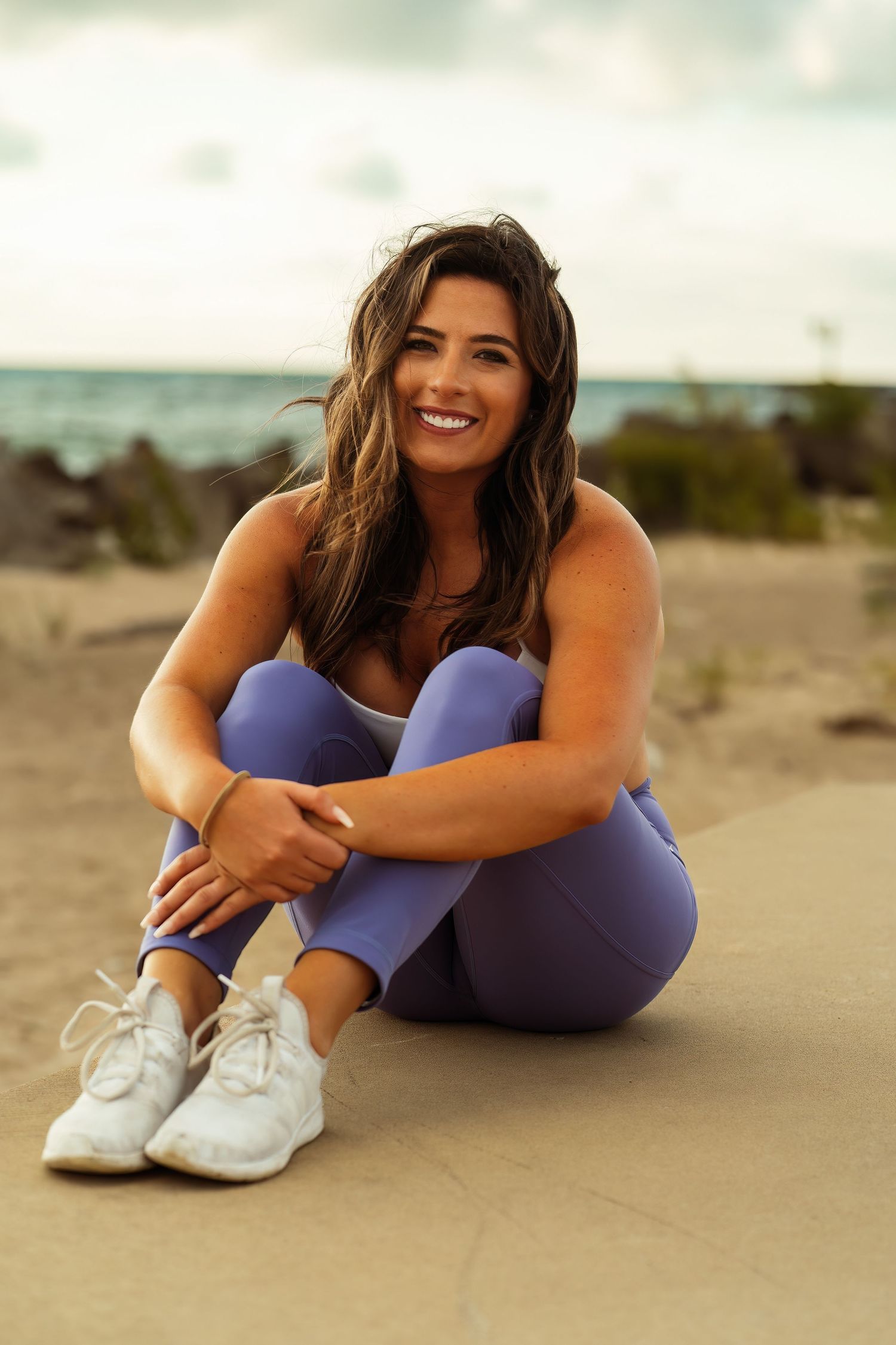Athletic woman in purple workout attire smiling while sitting on sandy beach.
