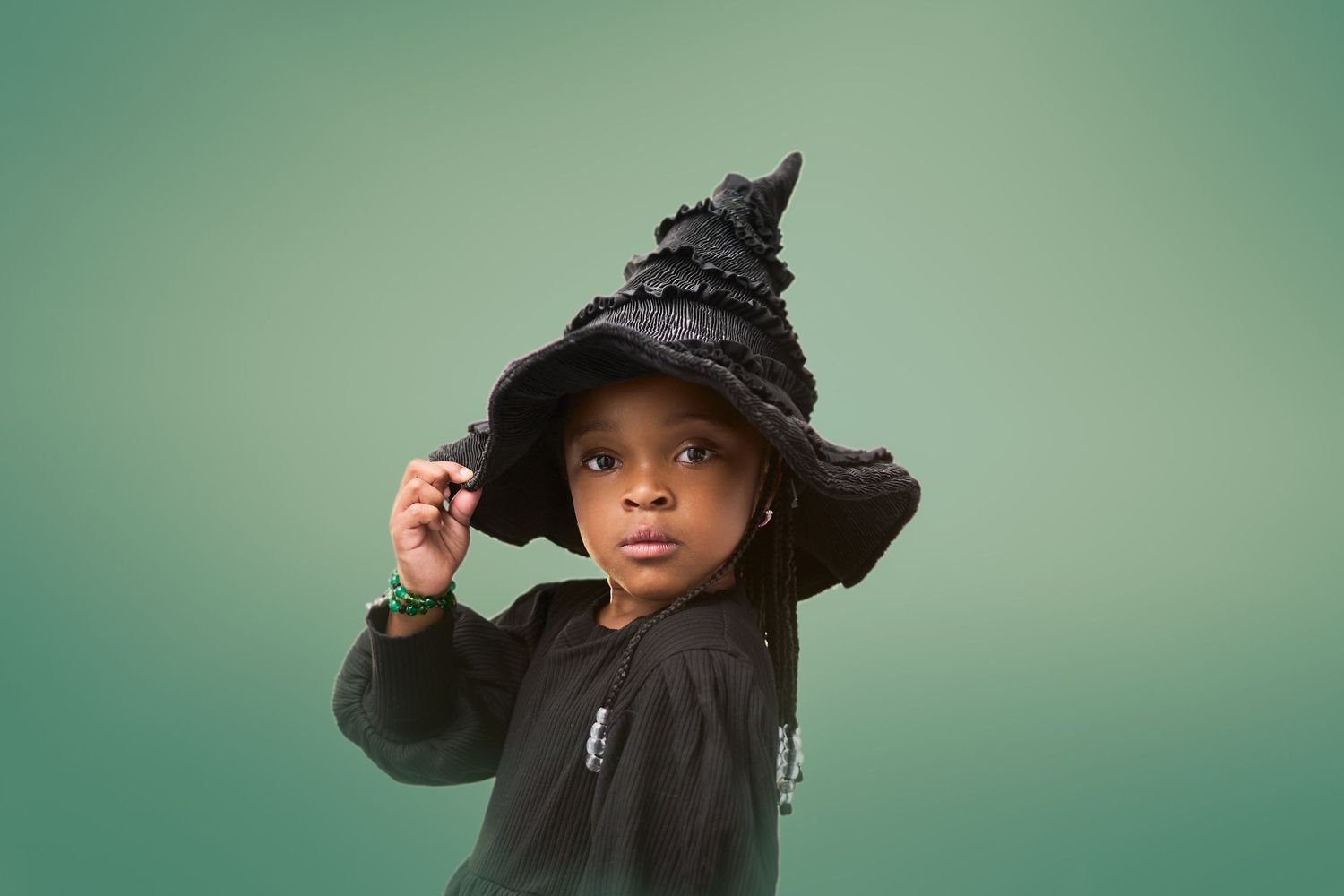 A child wearing a black witch hat and sweater poses against a teal background.