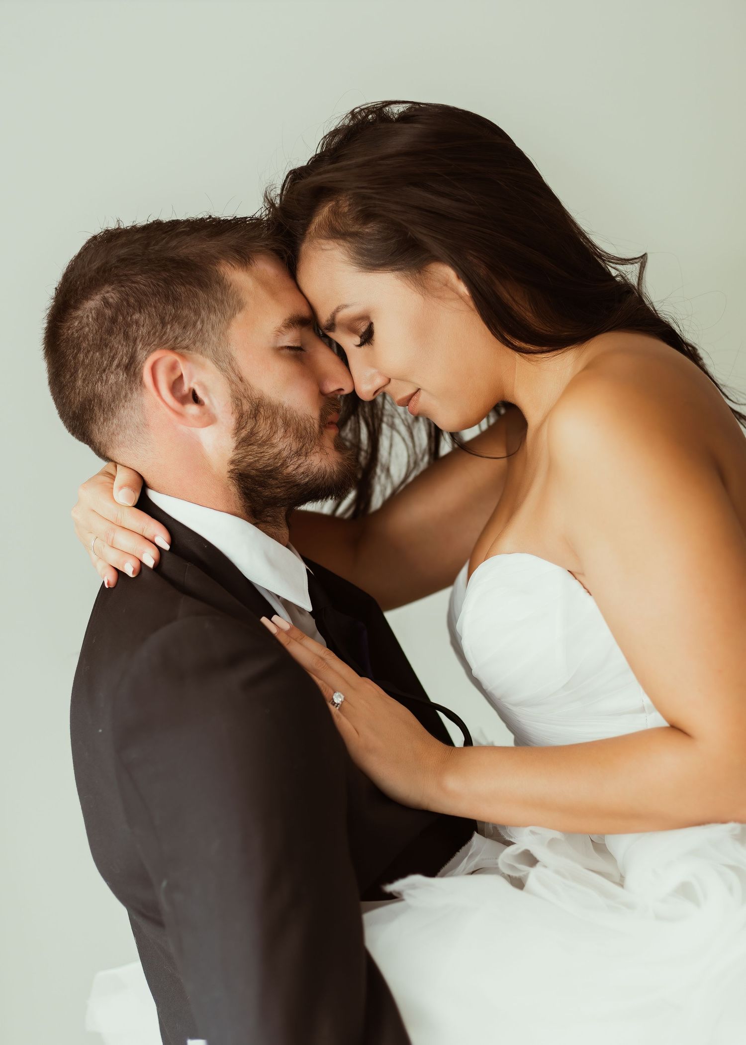 Intimate wedding portrait with couple in formal attire embracing against light background.