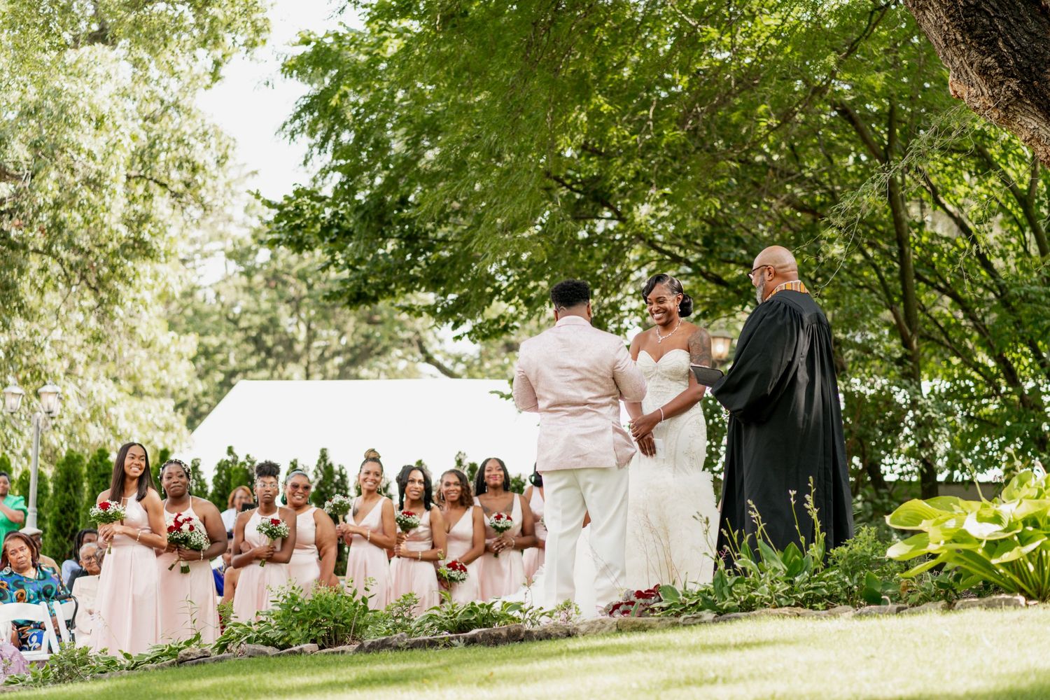 An outdoor wedding ceremony with bridal party in pink dresses watching from garden seating.