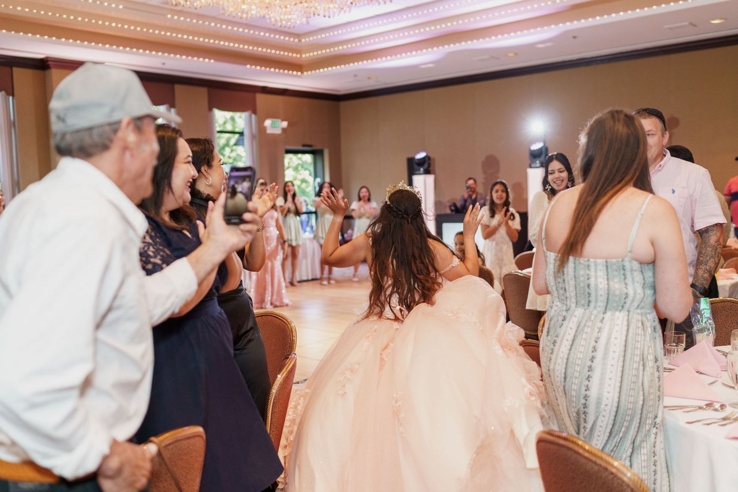 Guests capture photos and videos at a quinceanera celebration in a ballroom with a person in a peach gown at the center.