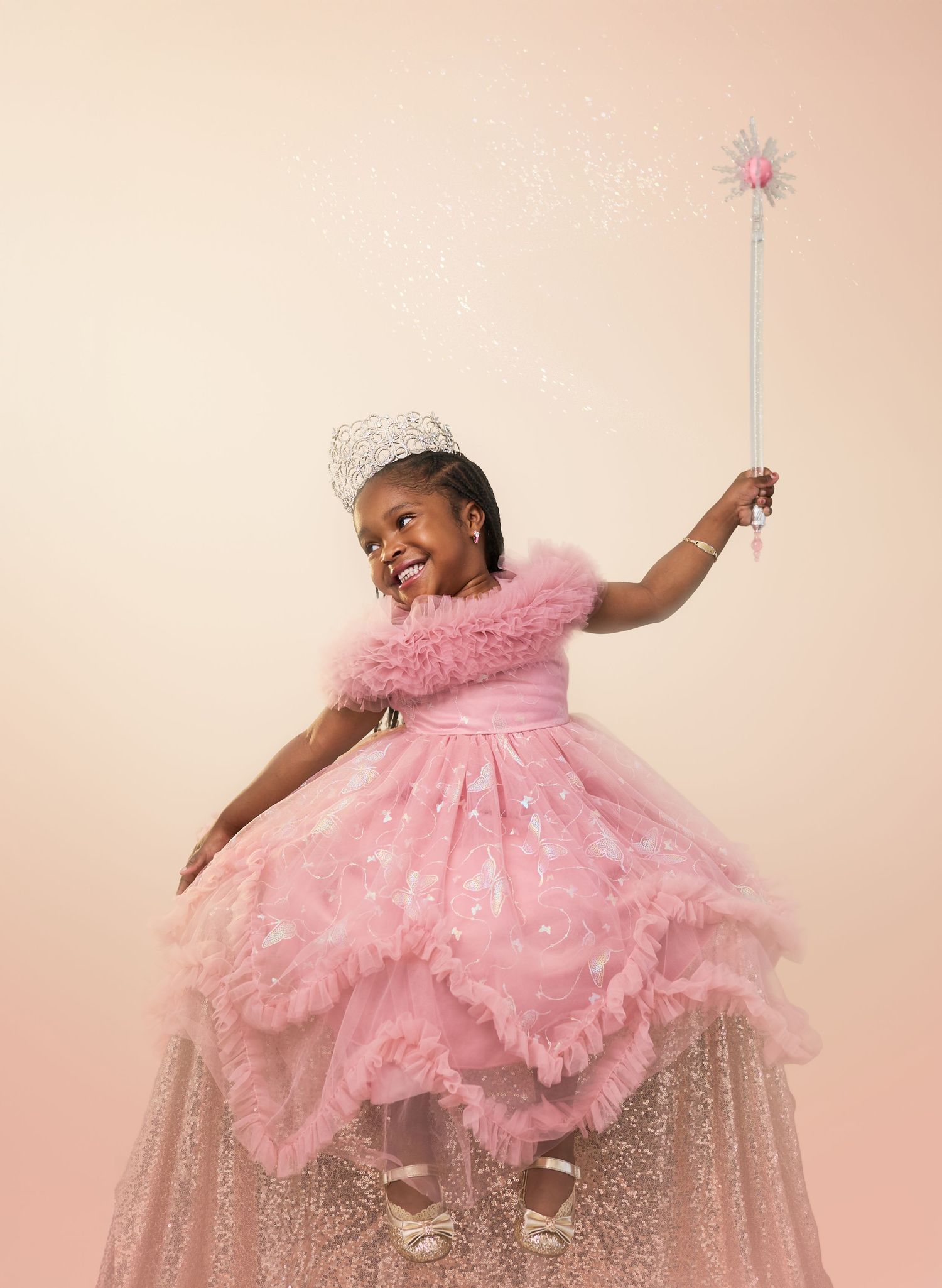 A child in a fluffy pink princess dress and crown twirls with a sparkly wand against a peachy pink background.