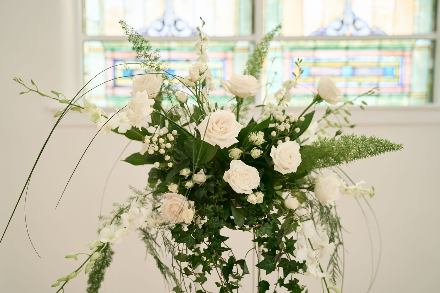 An elegant white floral arrangement with roses and greenery displayed against stained glass windows.