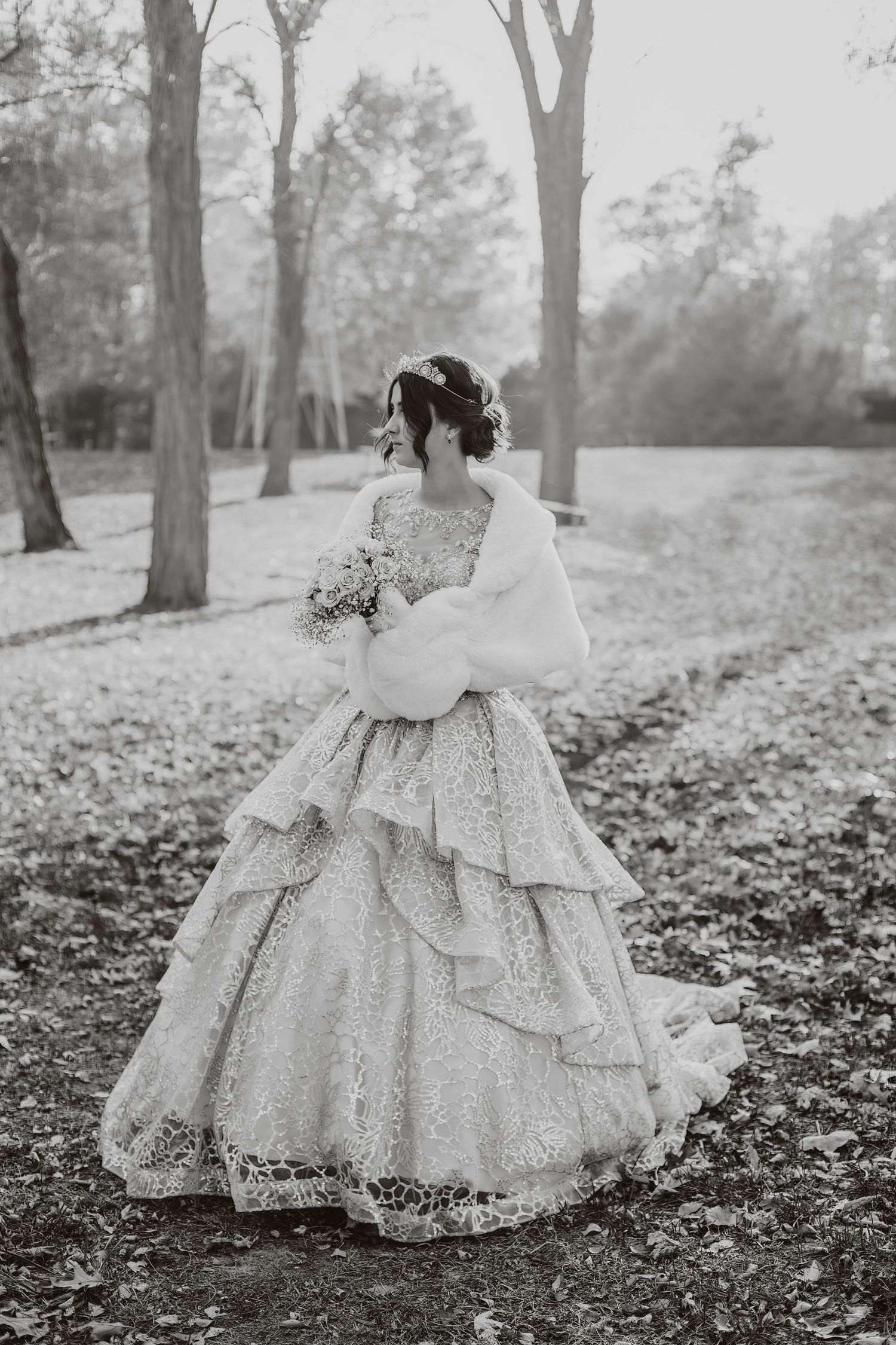 Black and white side view of bride in lace gown with fur wrap holding bouquet.
