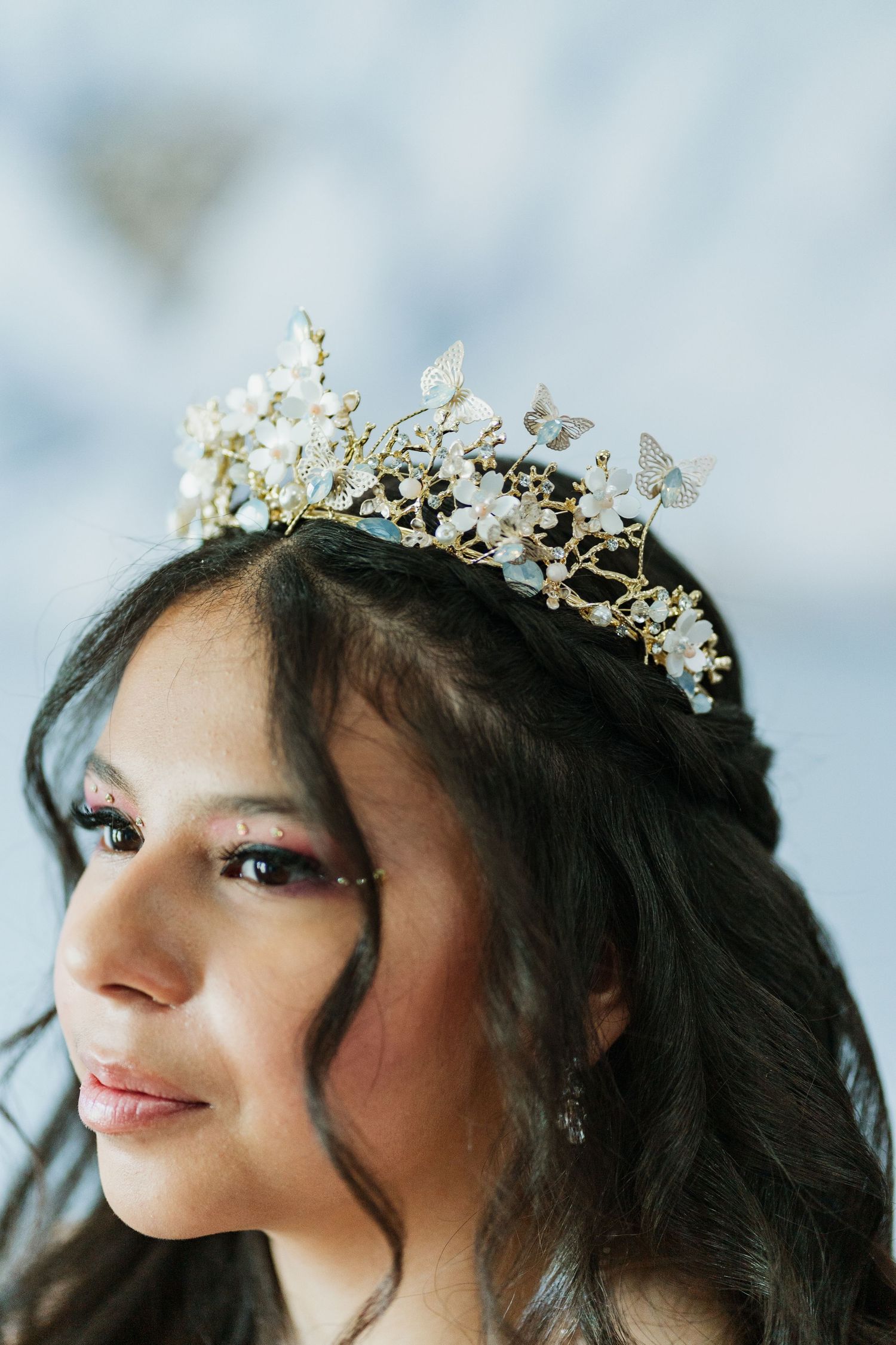 A person wearing an ornate gold crown with pearls and crystals gazes upward with a gentle smile against a light background.