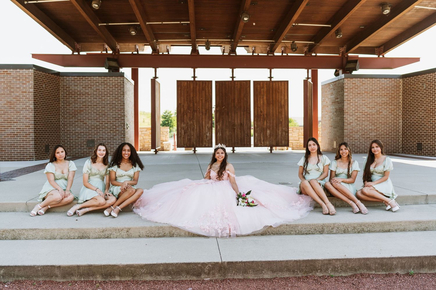 Wedding party members pose casually on concrete steps under a wooden pergola.
