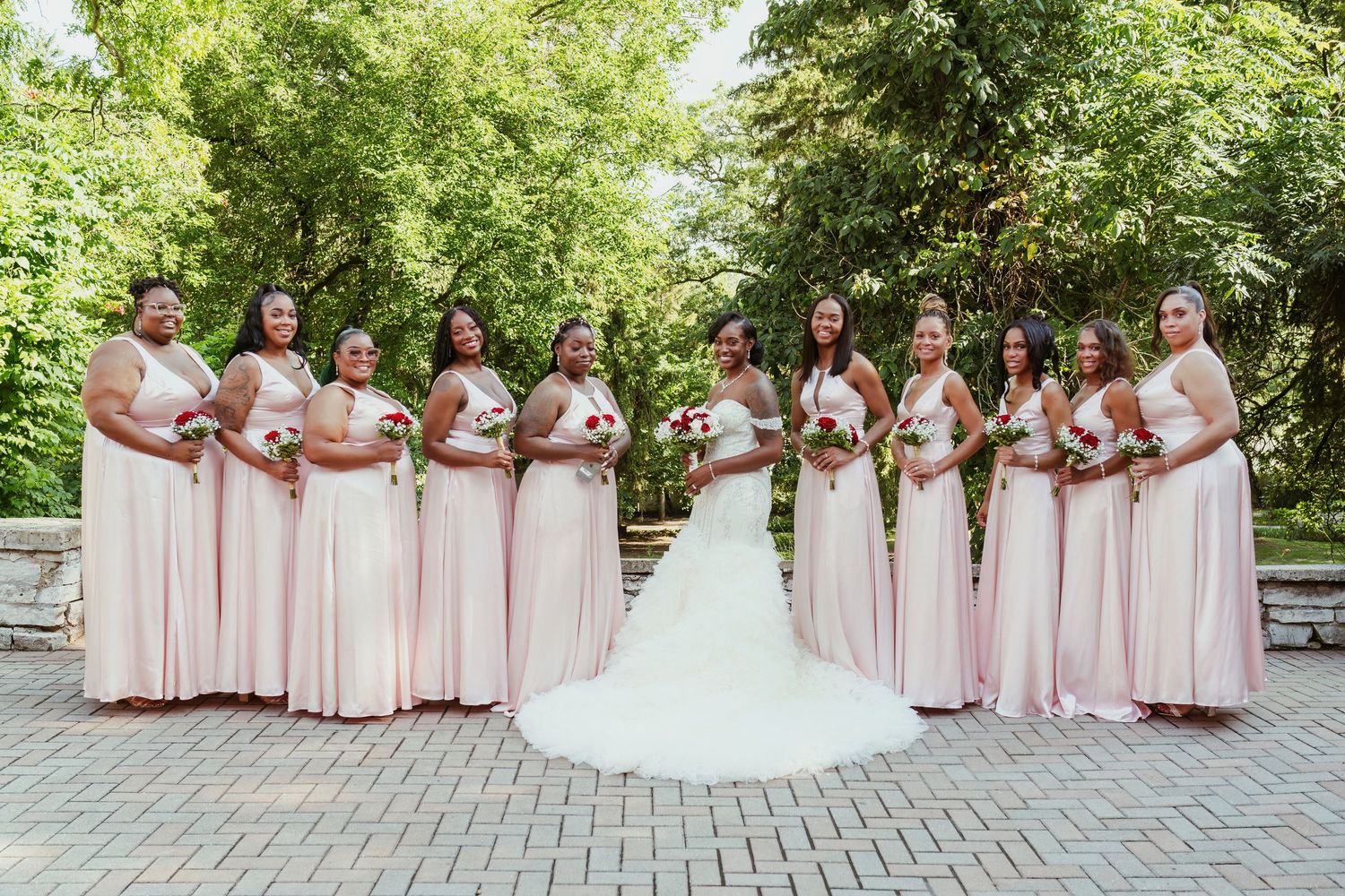 Bridesmaids in blush pink dresses stand with bride in white gown on brick pathway.