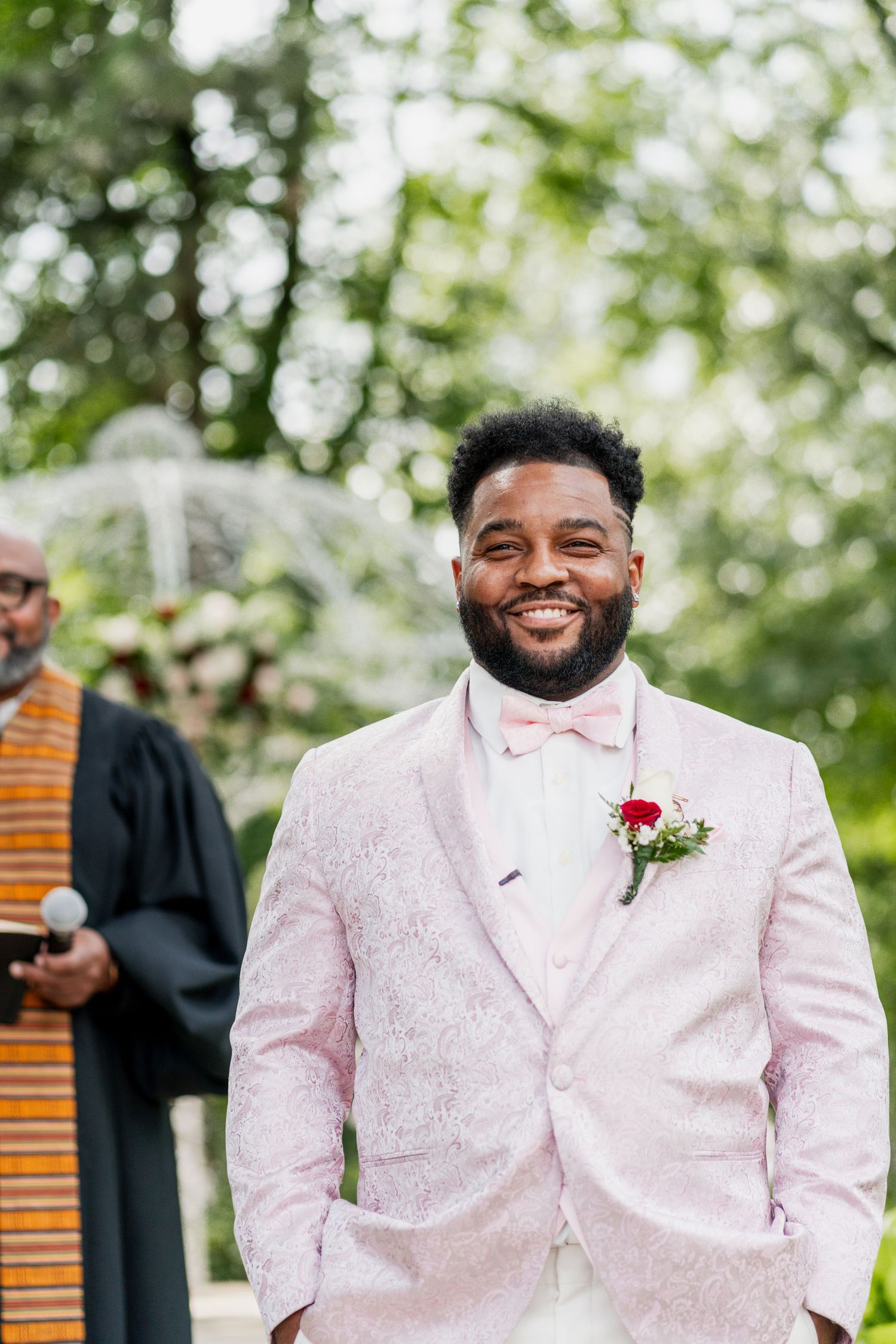 A joyful person wearing a light pink suit with a bow tie and boutonniere stands at an outdoor wedding ceremony.