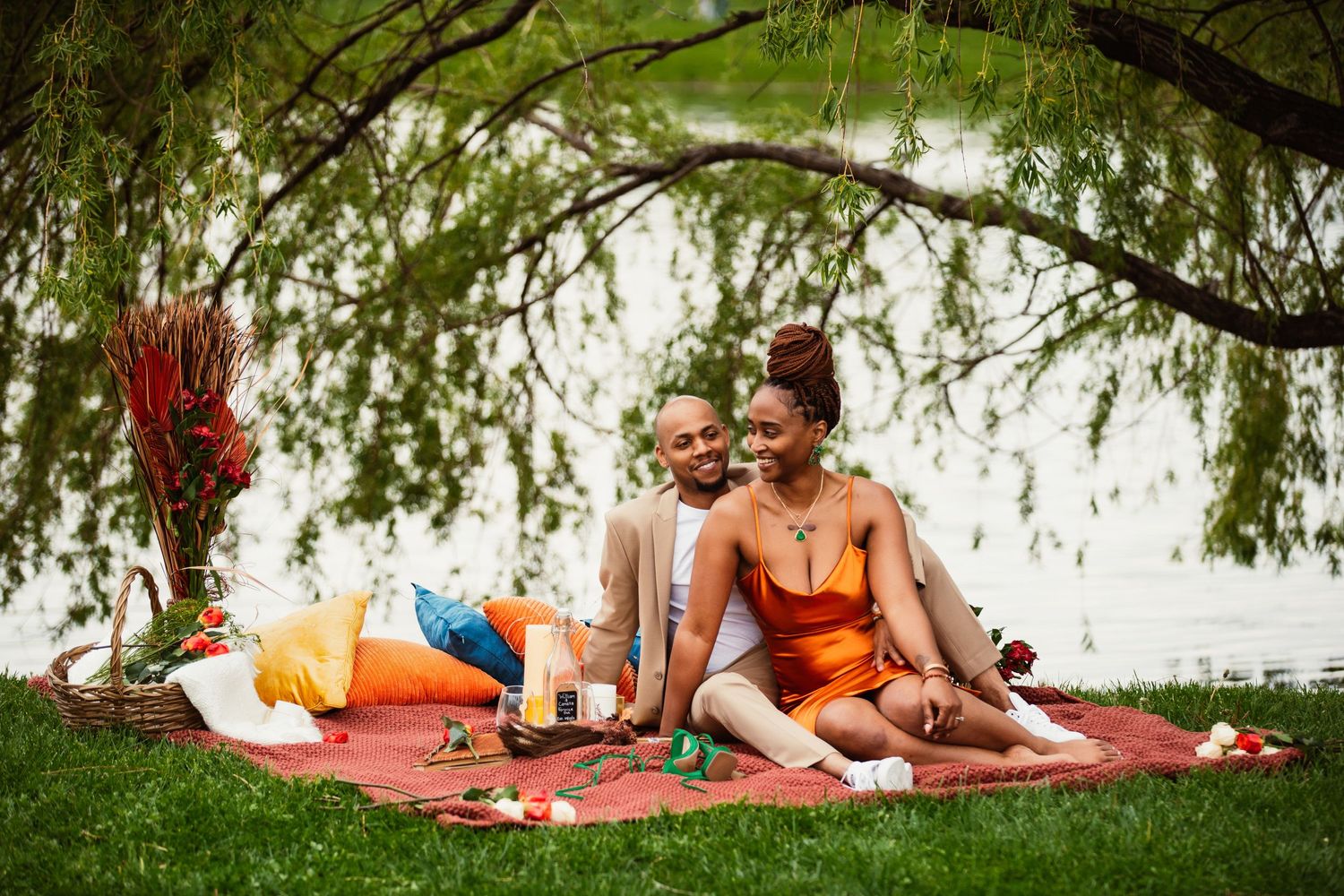 Romantic outdoor picnic setup with couple lounging under willow tree branches.