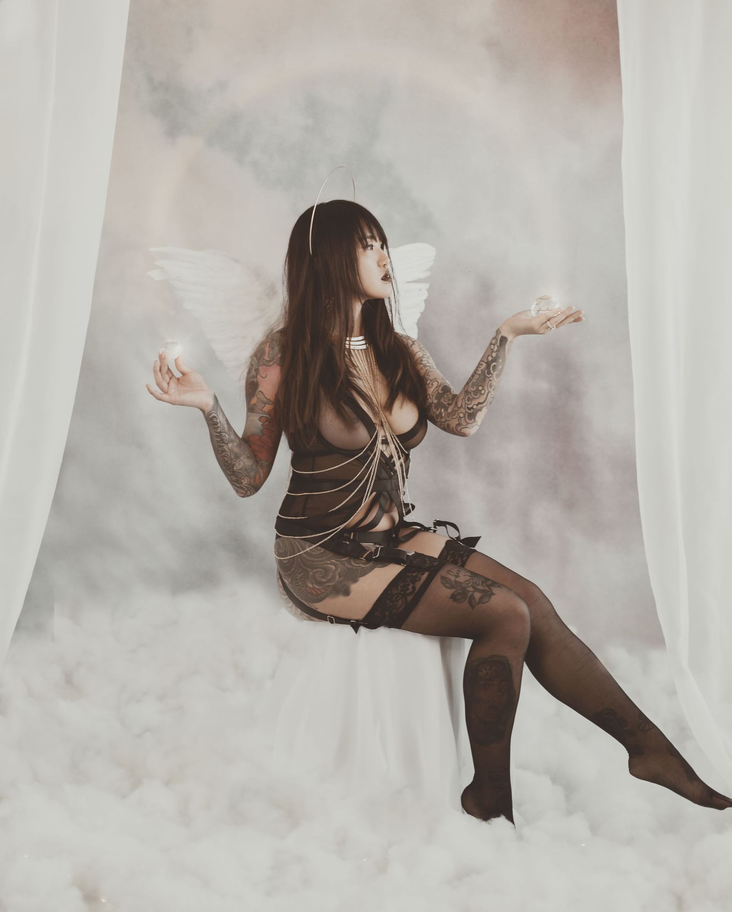 Ethereal angel-themed boudoir photo with see-through dress and wings in misty setting.