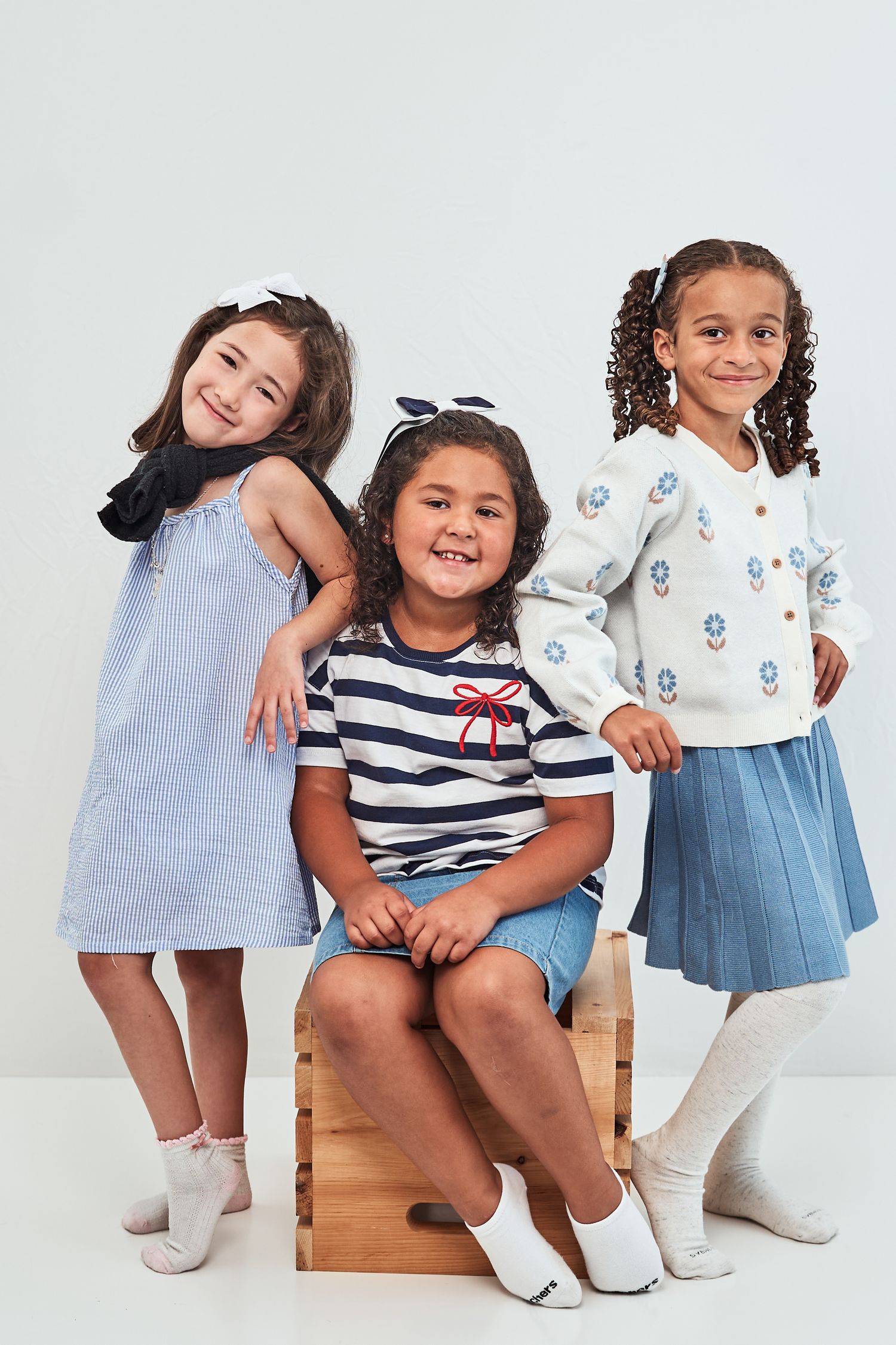 Three cheerful children in casual spring outfits pose together on a wooden box against a white background.
