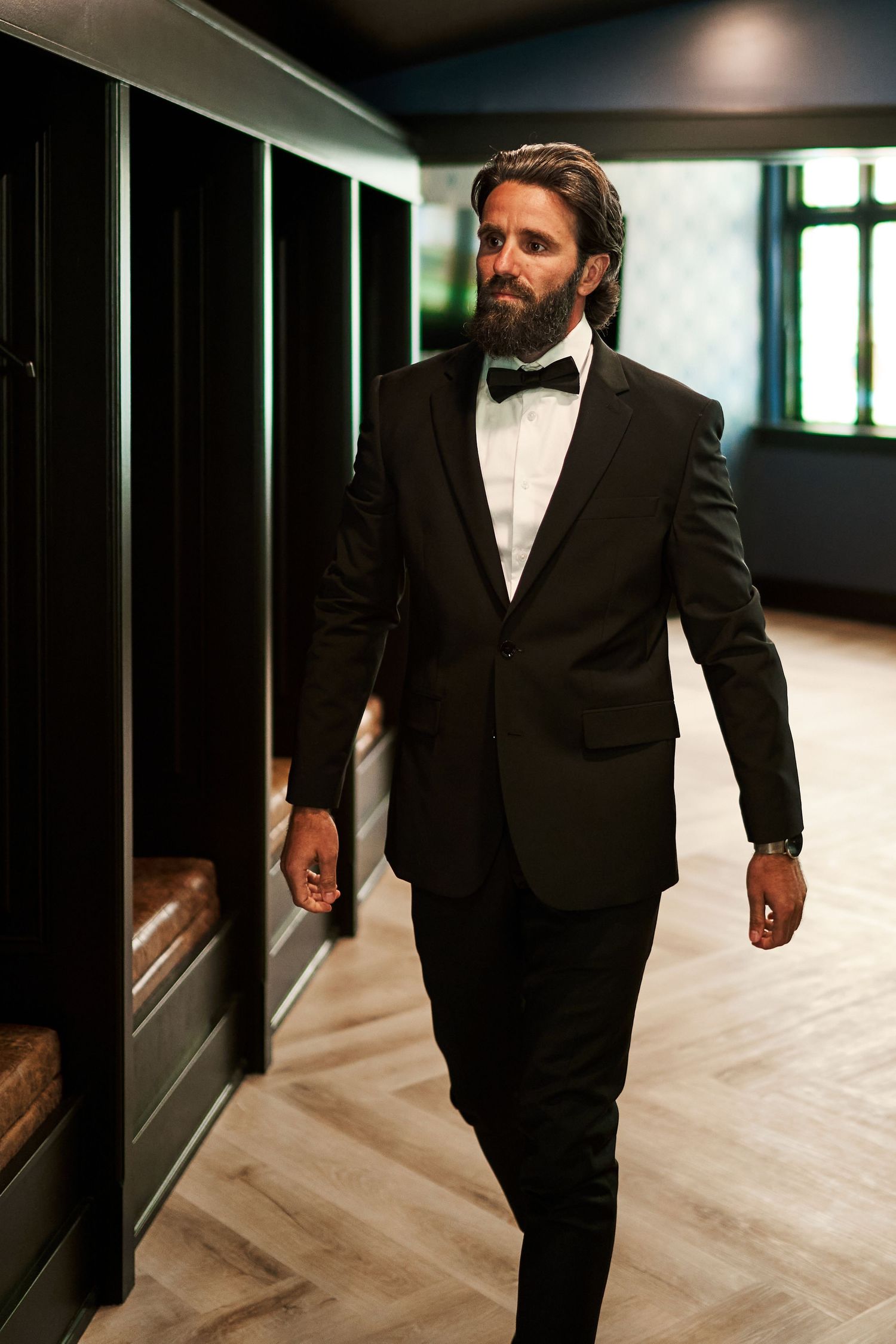 A bearded gentleman in a classic black tuxedo and bow tie walks through an elegant hallway with wooden floors.