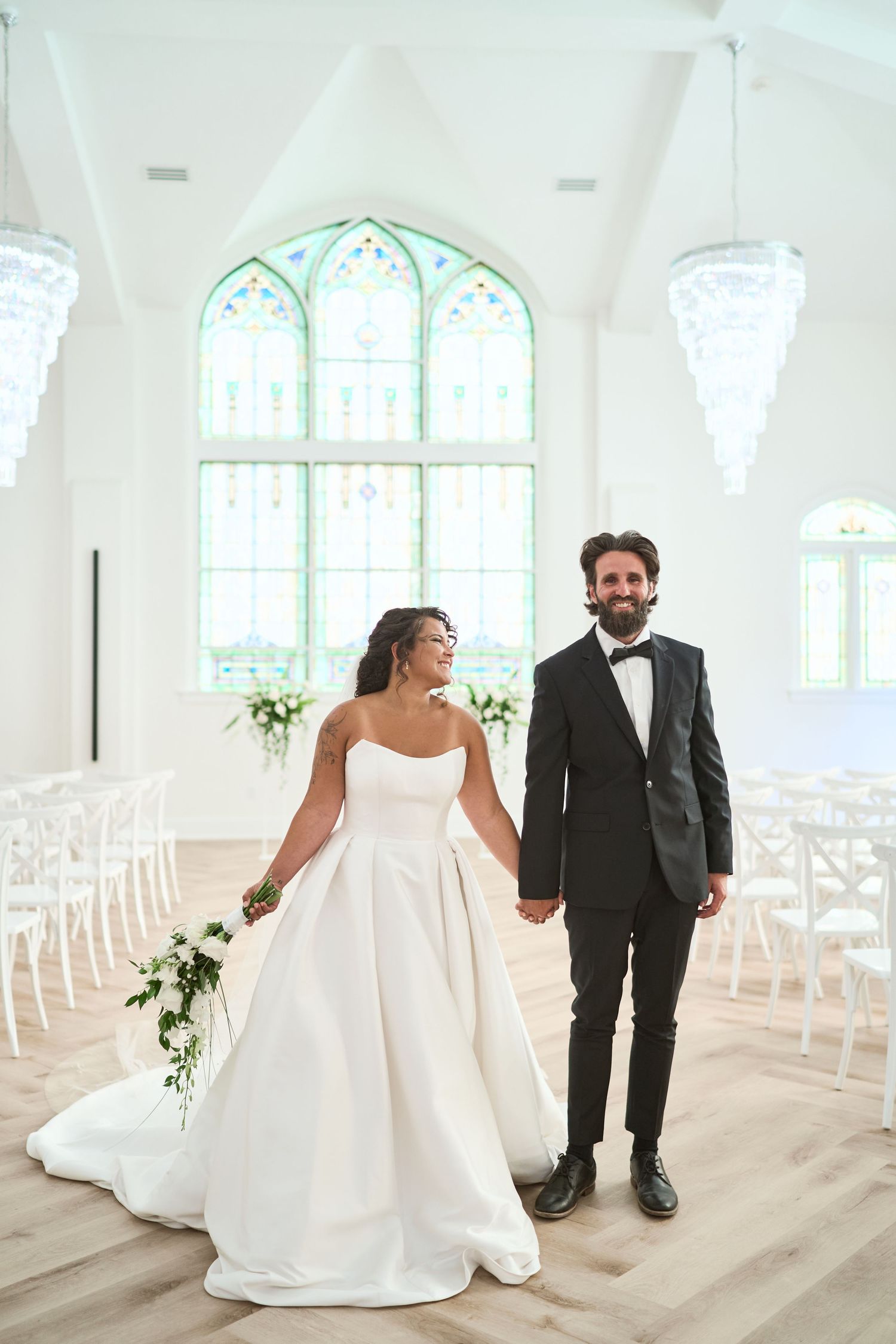 A bride in a flowing white gown walks with her groom in a bright, sunlit chapel with stained glass windows.