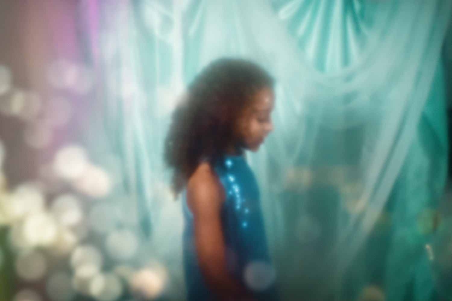 A young girl in a blue sequin dress stands in a dreamy, bokeh-lit teal backdrop setting.