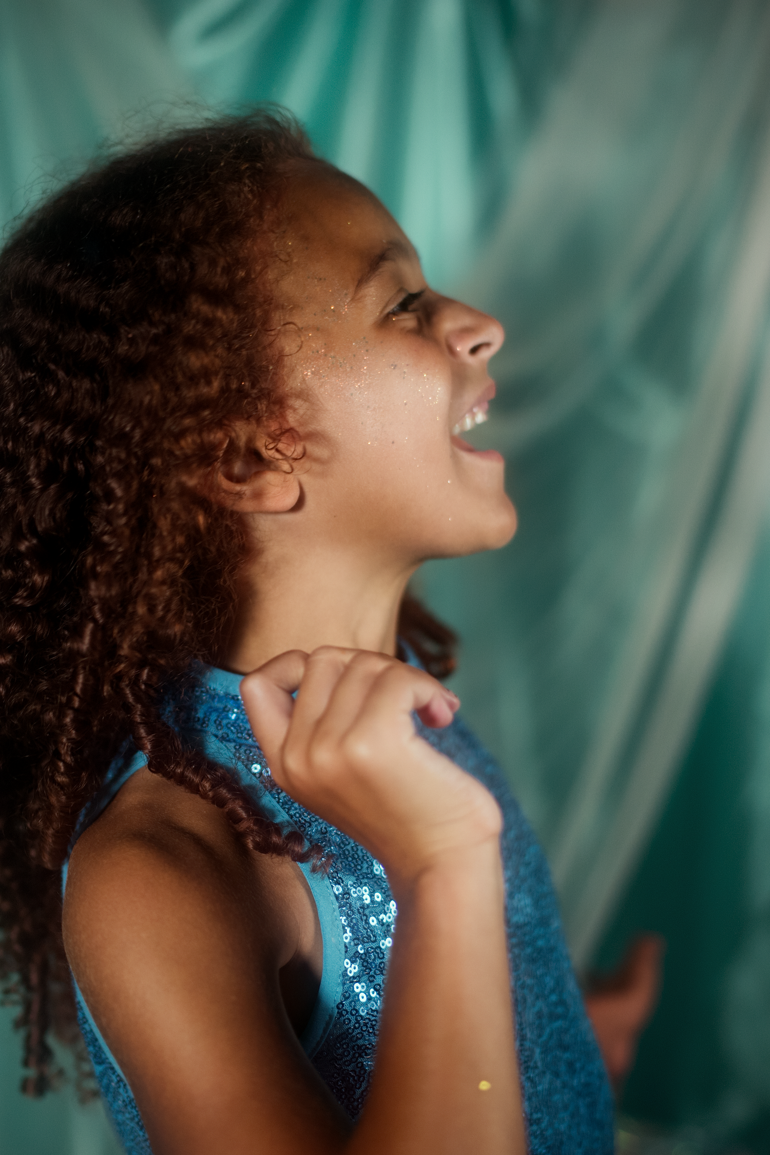 A joyful young girl with curly hair laughs in a blue sequin dress against a teal backdrop.